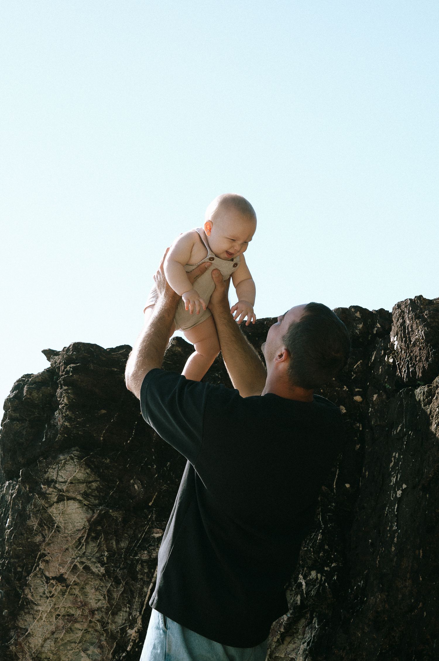 A parent lifts a joyful baby up into the air against a bright sky.