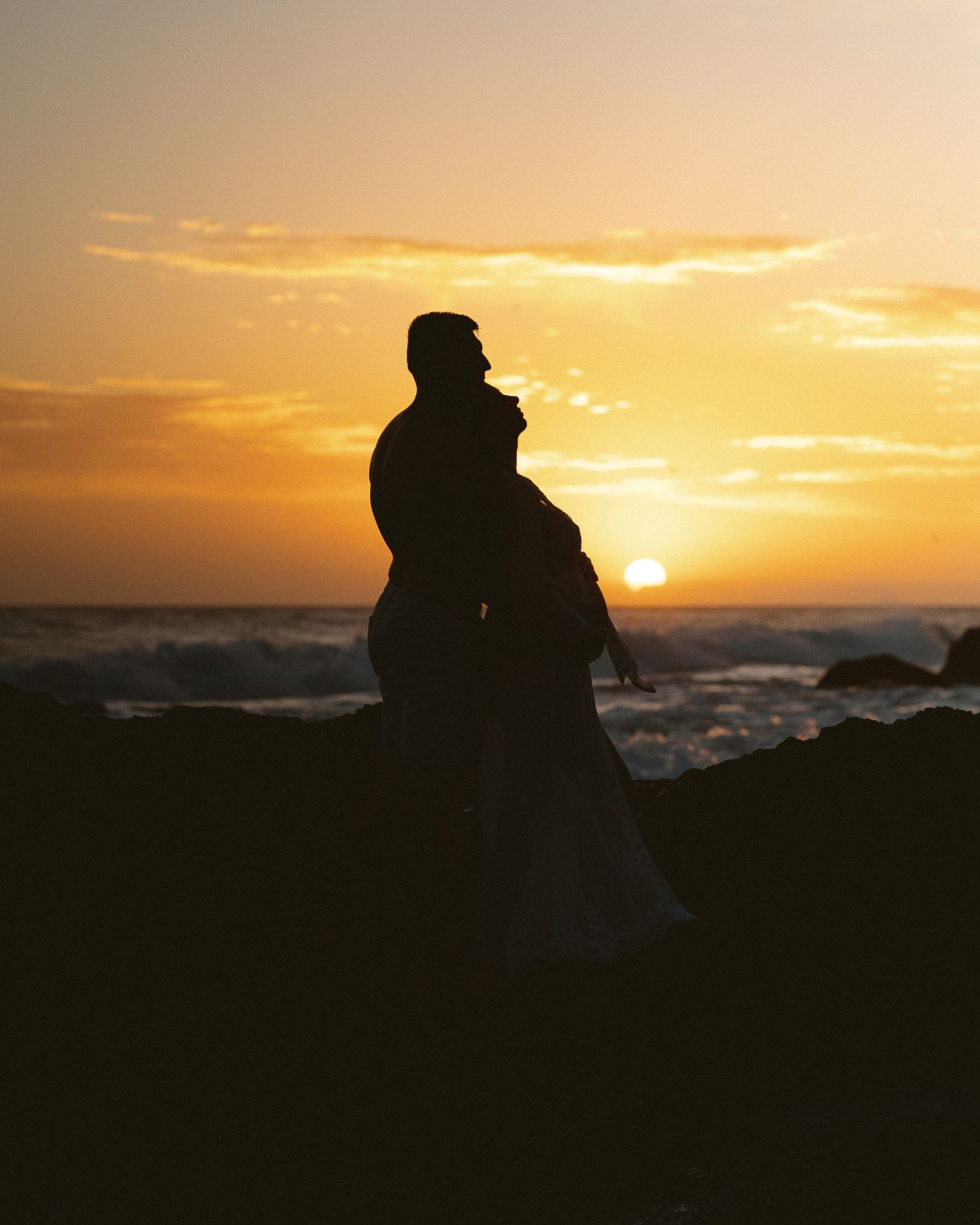 Silhouette of a couple embracing on rocky shore at golden sunset with ocean waves in background.