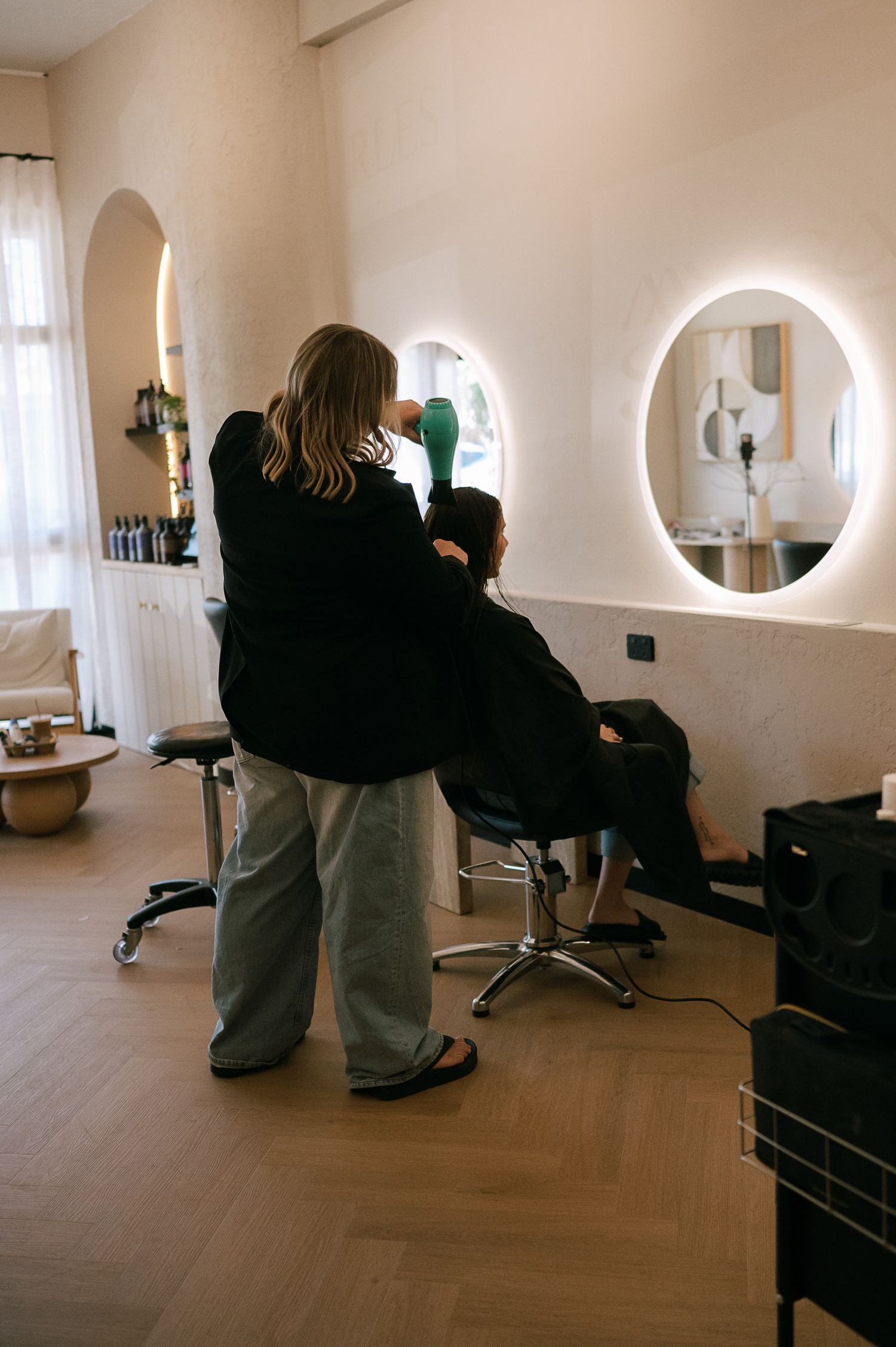 Hairstylist blow-drying client hair in modern salon with round LED mirrors and warm white interior.