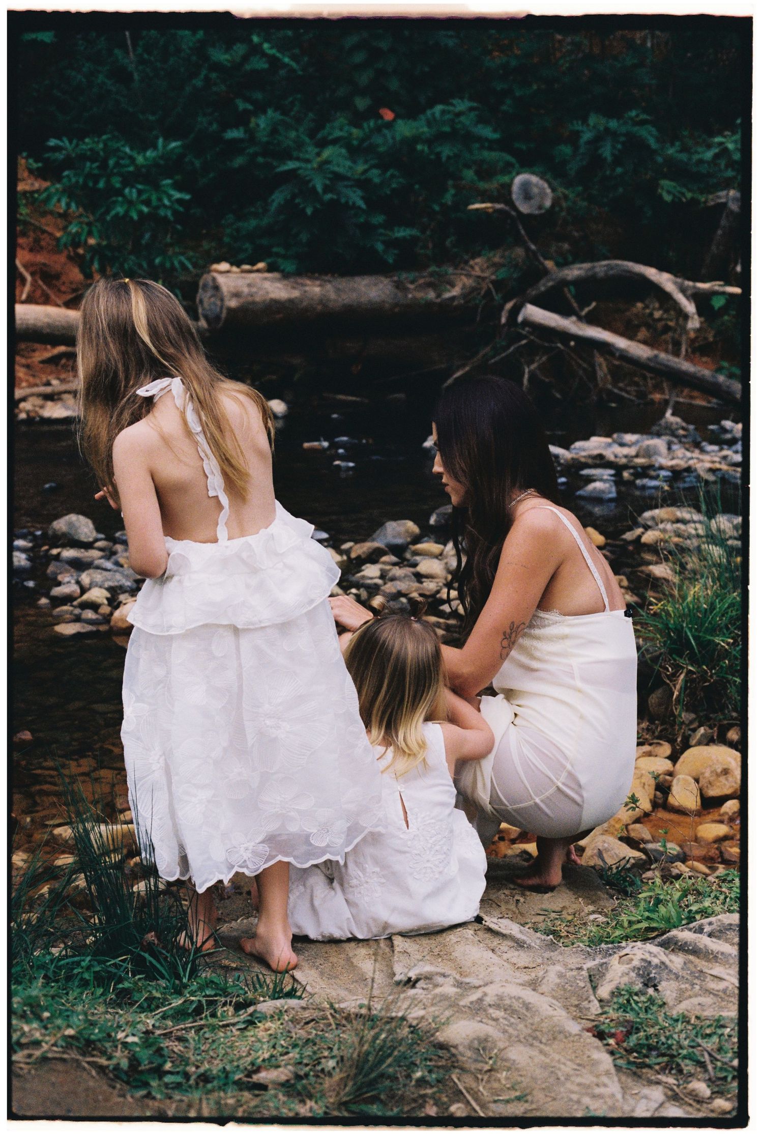 Three females in white dresses crouch by a rocky stream surrounded by lush greenery.