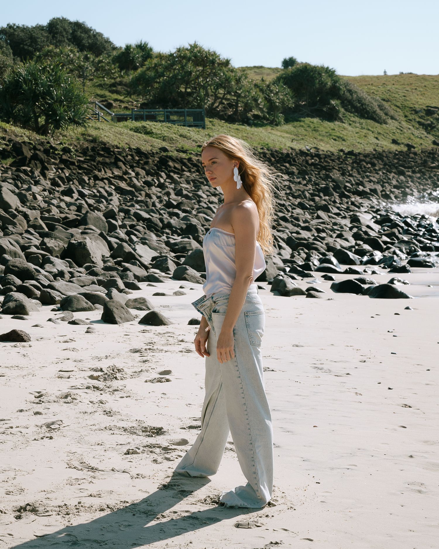 Woman in white strapless top and wide-leg pants walking on a rocky beach with green hills in the background.