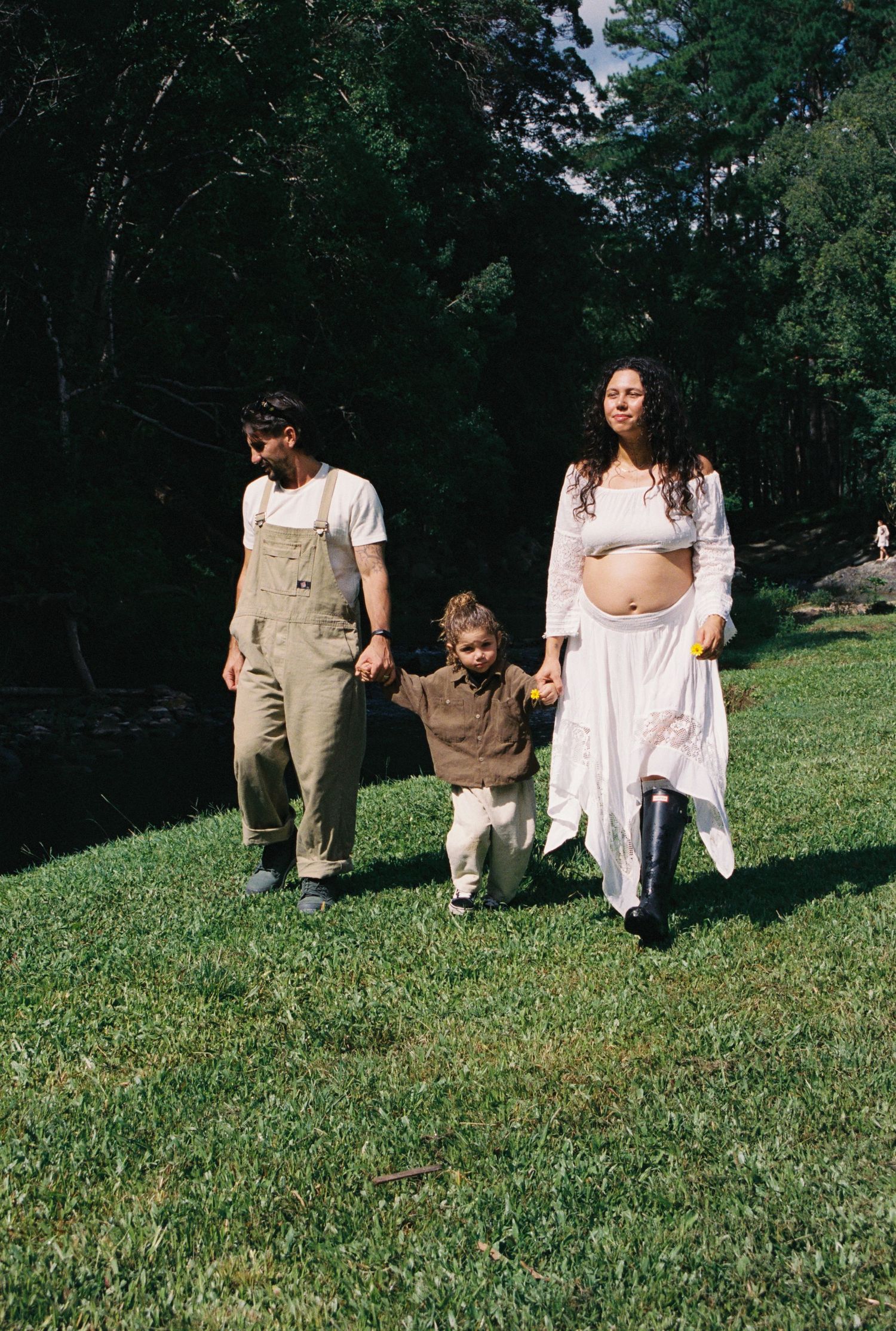 Pregnant woman in white dress walks with man in overalls and toddler on green grass near trees.