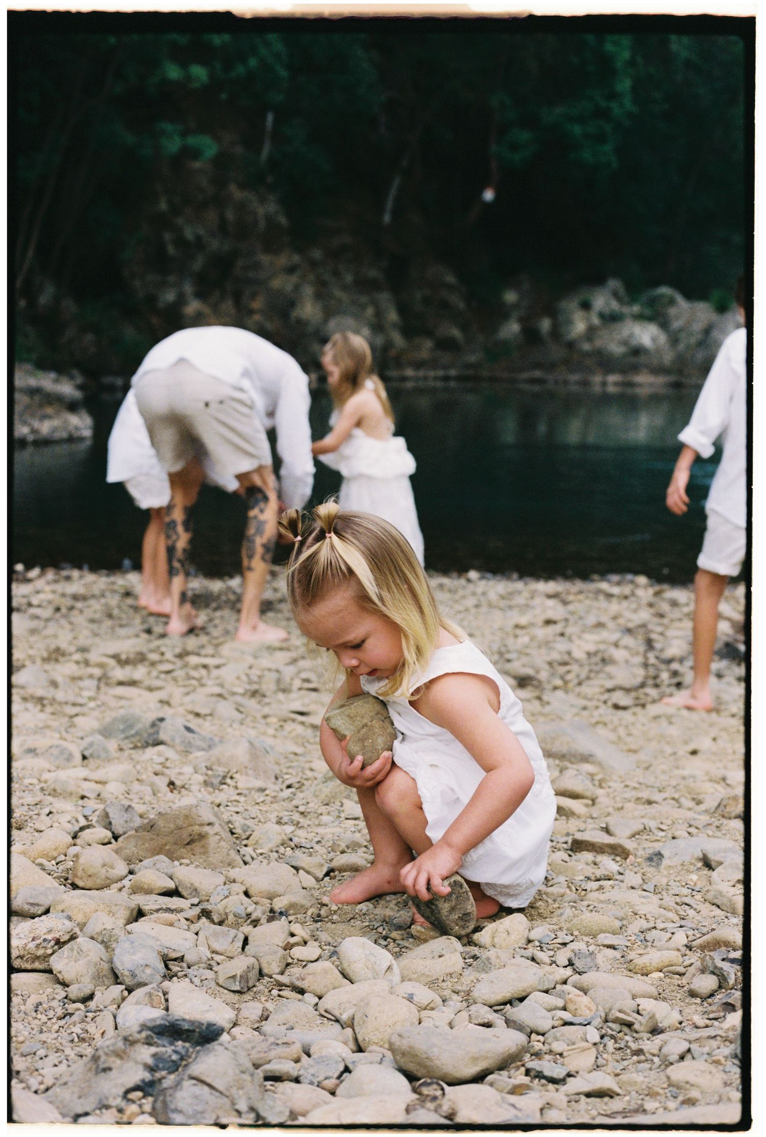Young blonde girl in white outfit stacking rocks on a pebble beach while family explores the shoreline.