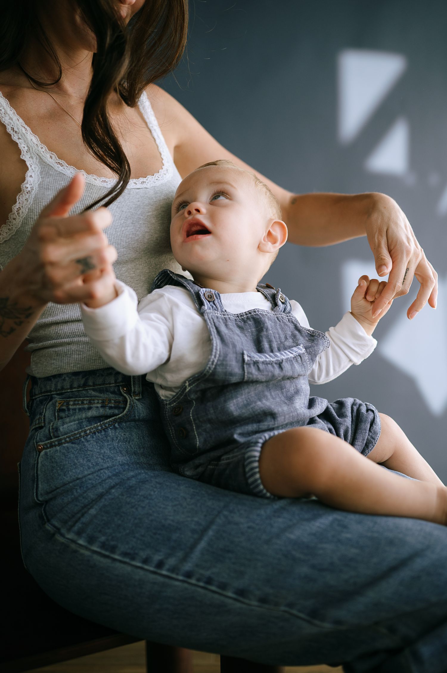 A mother holds her happy baby in denim overalls on her lap, both enjoying a playful moment together.