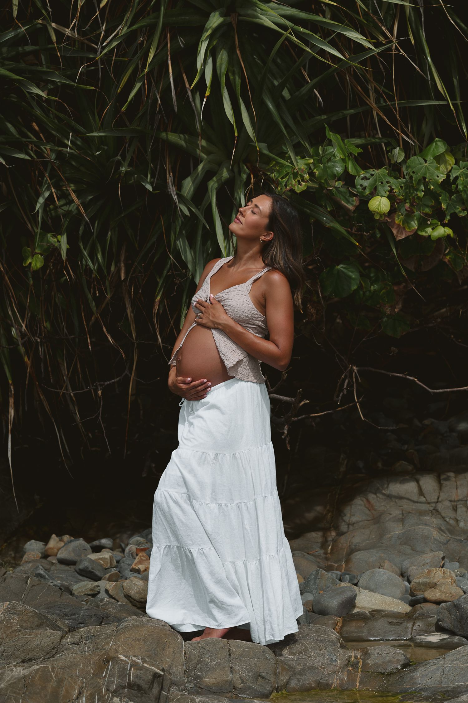 Outdoor maternity photoshoot featuring white dress against tropical foliage backdrop.