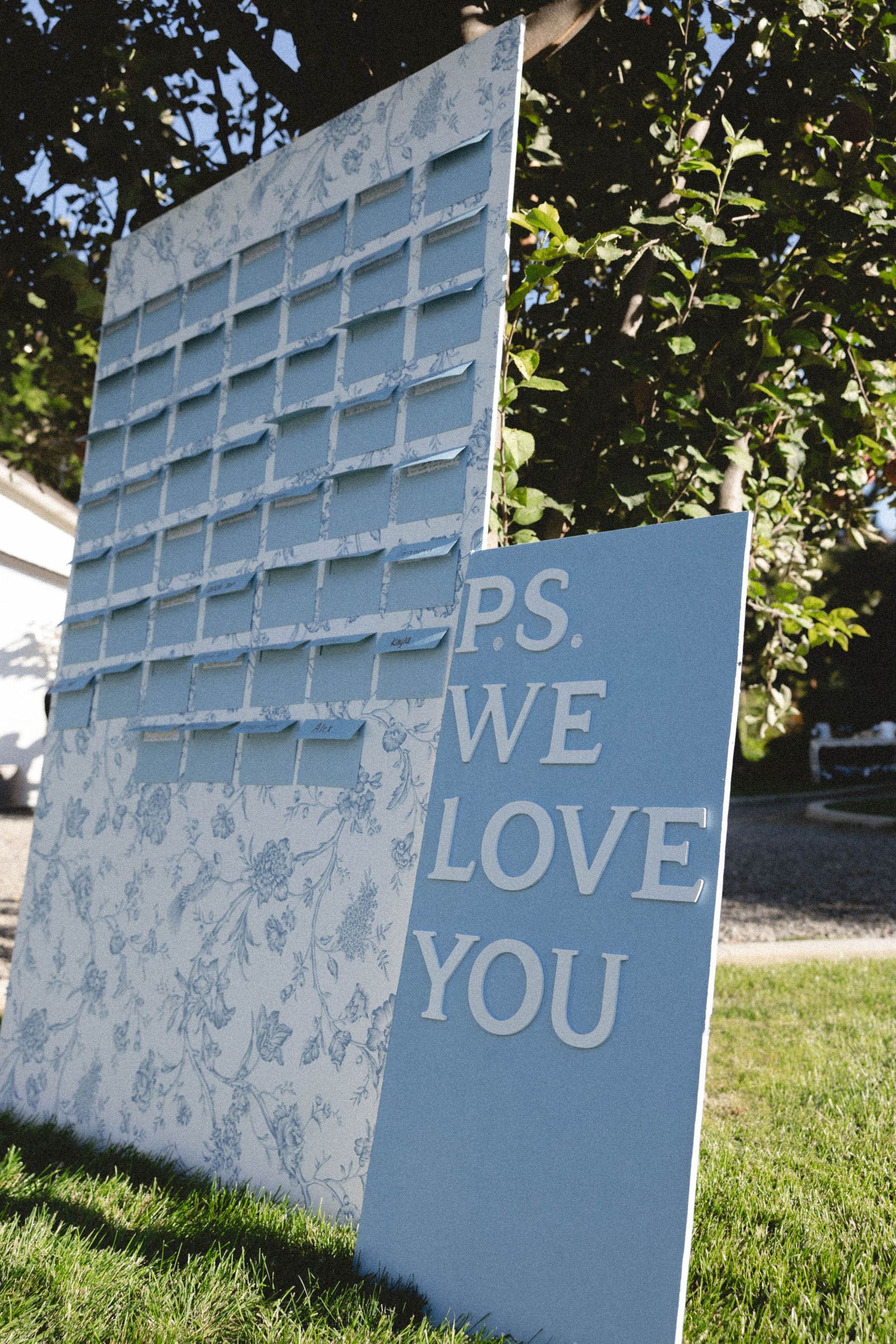 Light blue and white outdoor display panel with text reading 'PS, WE LOVE YOU' against green lawn and foliage.