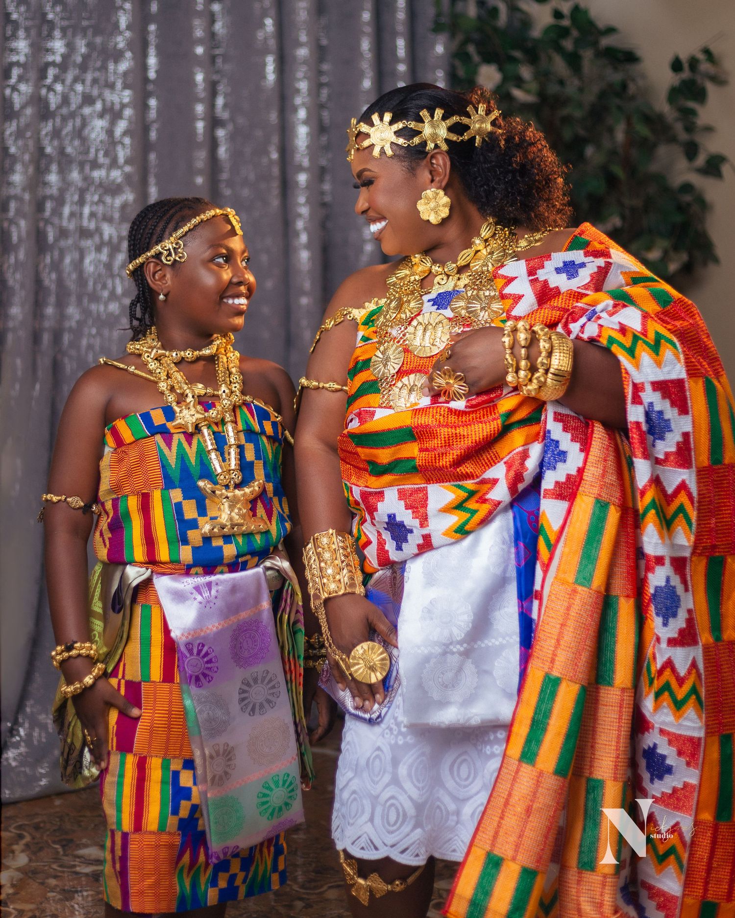 Traditional African clothing featuring vibrant kente cloth patterns in orange, red and gold with ornate jewelry and headpieces.