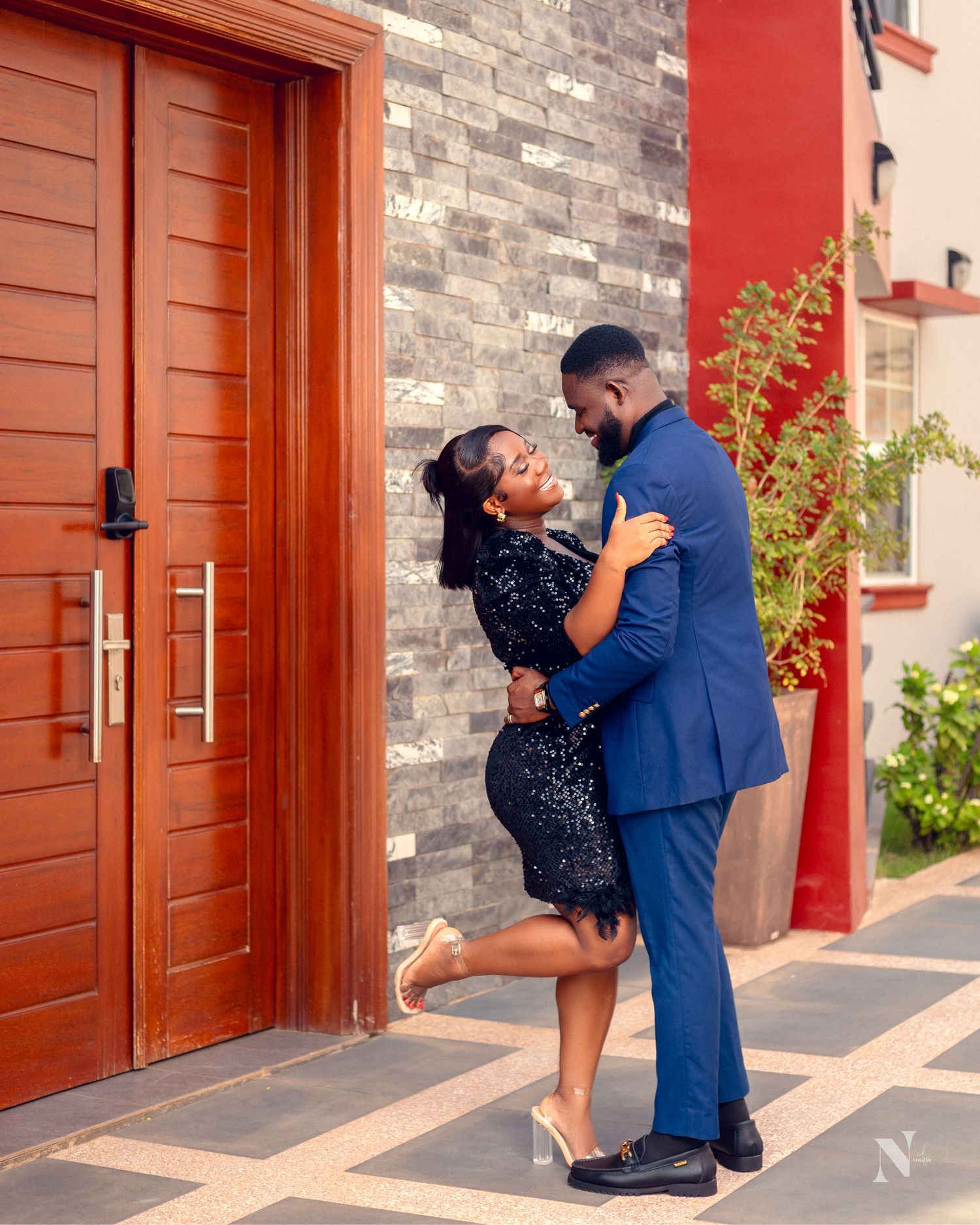 A couple shares a romantic moment outside a brick building with a red door, the woman in a polka dot dress.