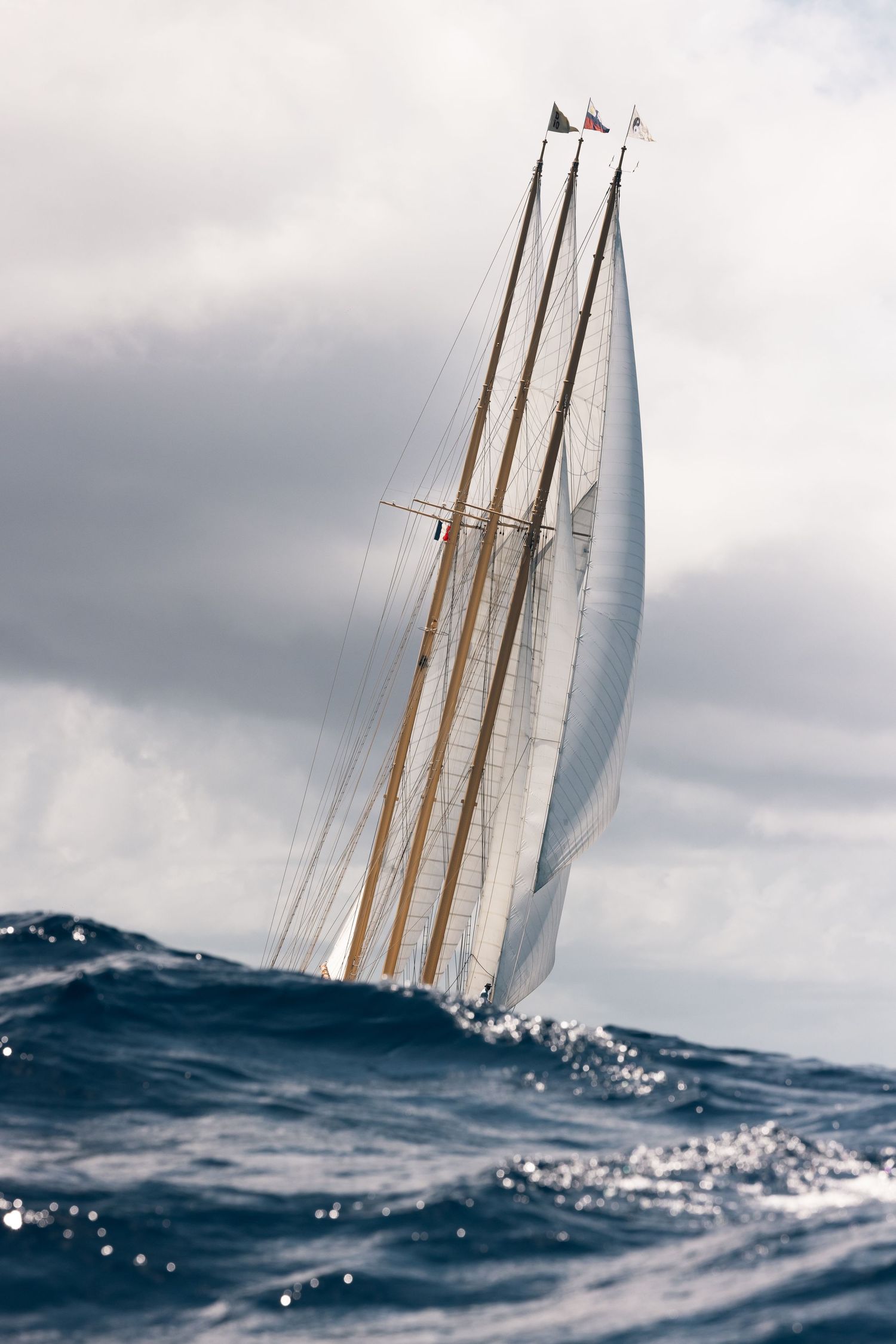A tall sailing ship with white sails navigates rough ocean waves under a dramatic cloudy sky.