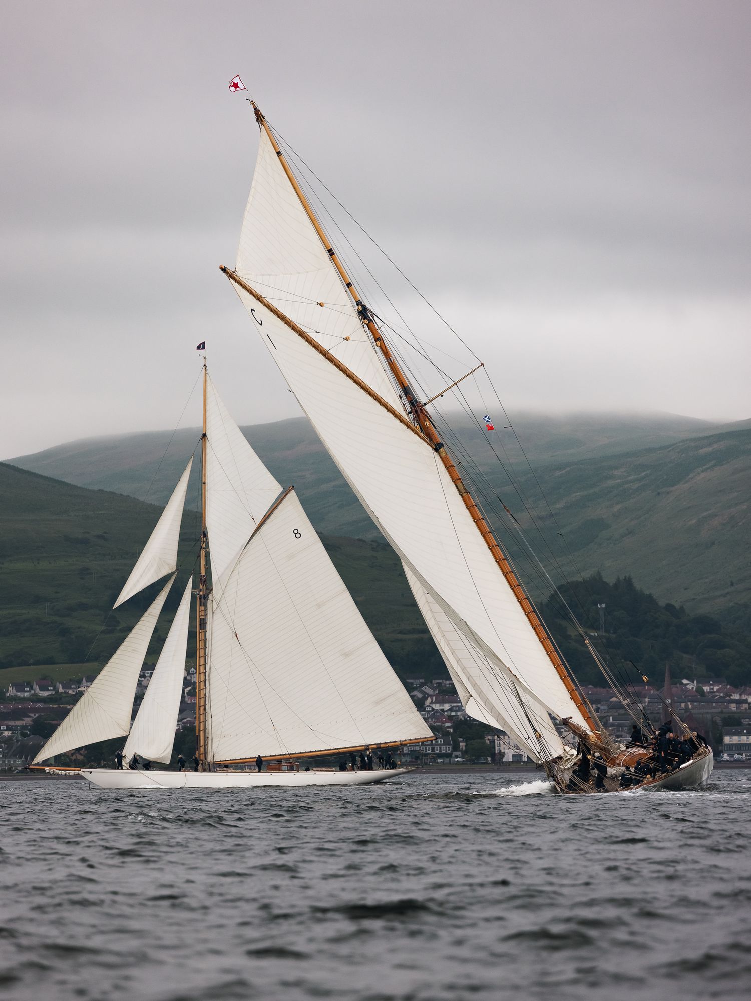 Two classic wooden sailing yachts with white sails racing on choppy waters with green hills in the background.