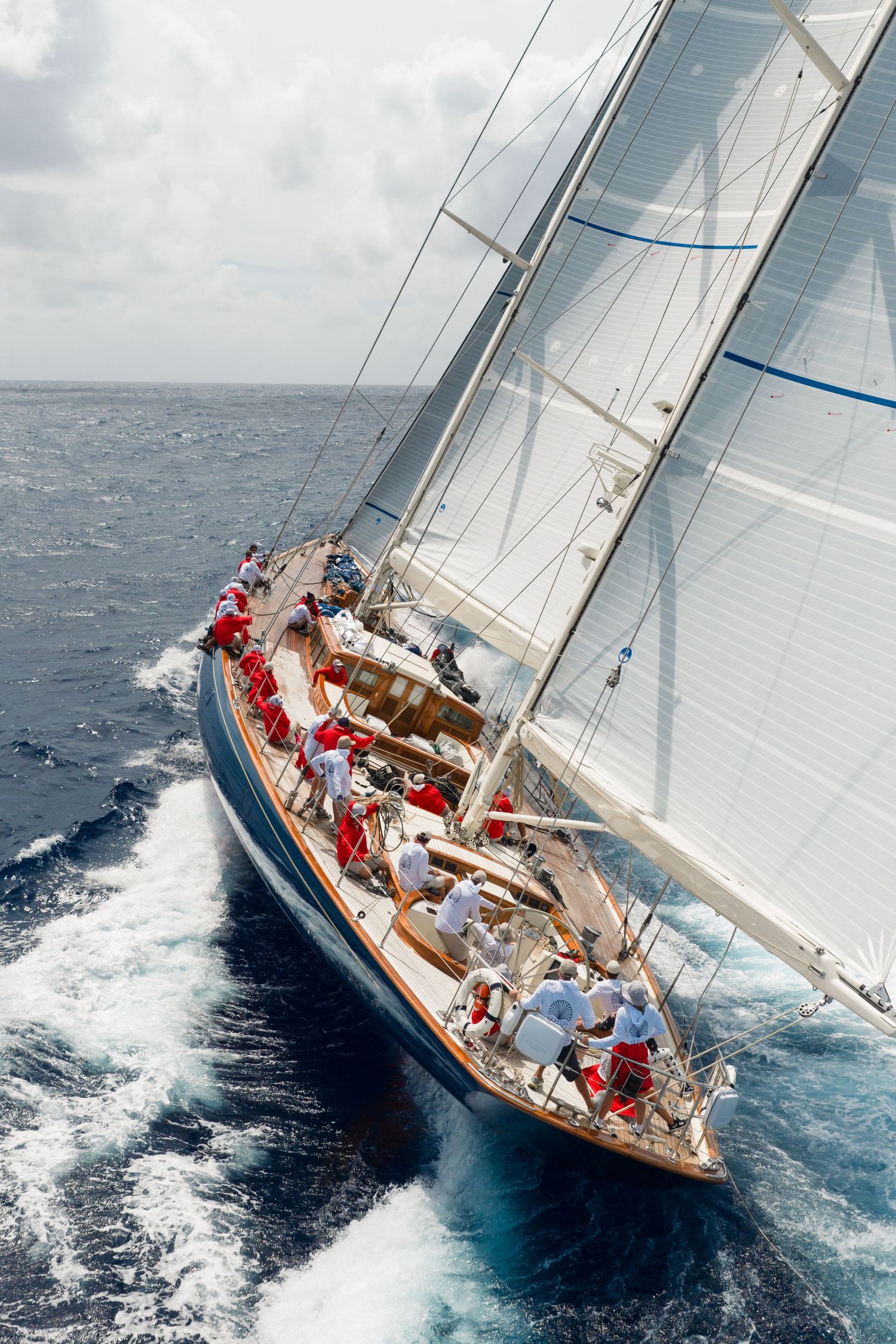 Aerial view of a large classic sailing yacht with white sails cutting through blue ocean waves with crew on deck.