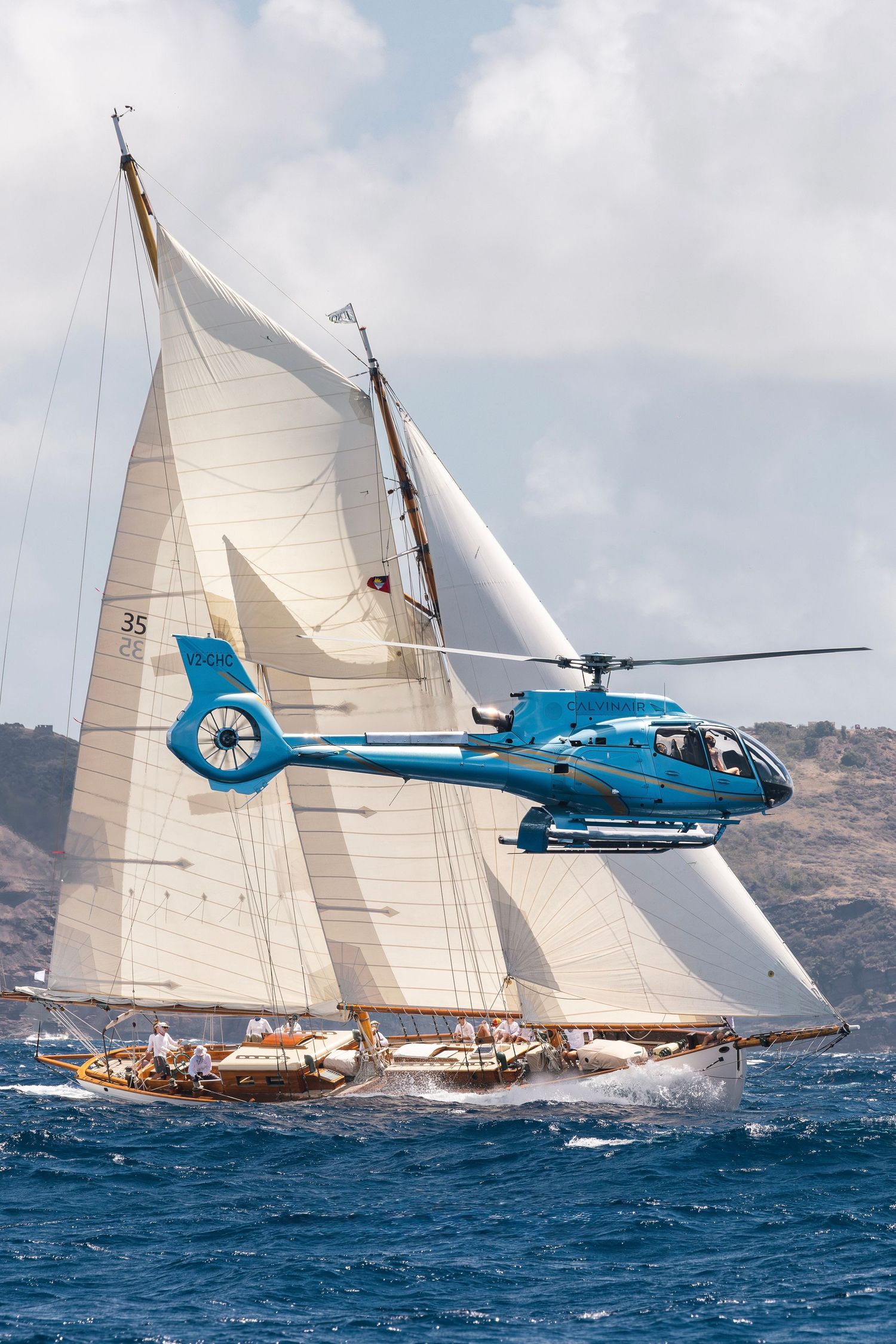Blue helicopter flying alongside a classic tall ship with white sails racing on choppy ocean waters under cloudy skies.