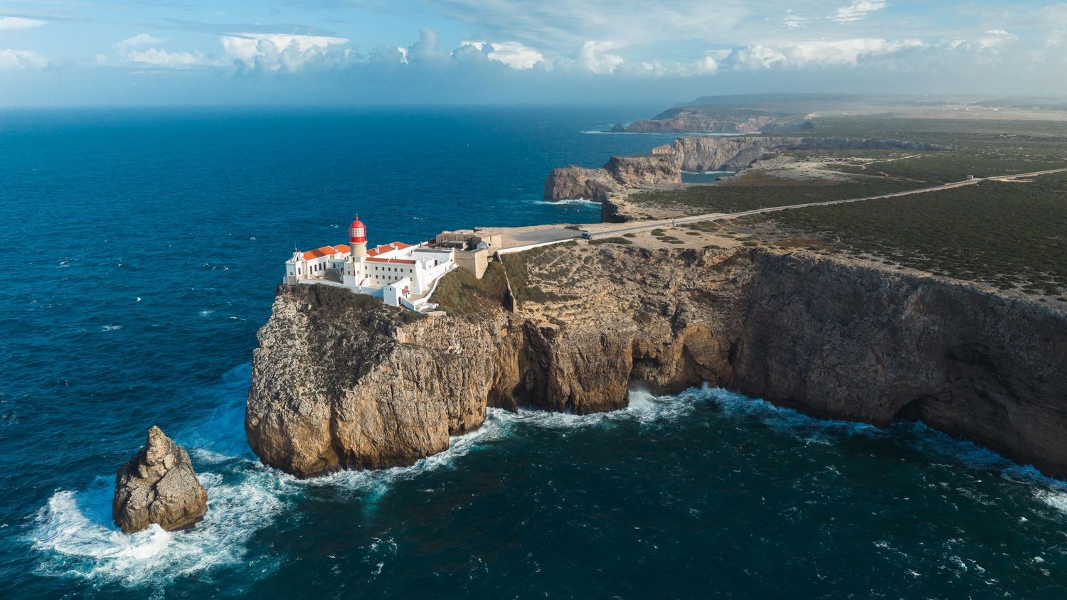 Aerial view of a lighthouse on dramatic rocky cliffs at Cabo de São Vicente, Portugal, with deep blue Atlantic Ocean waves.