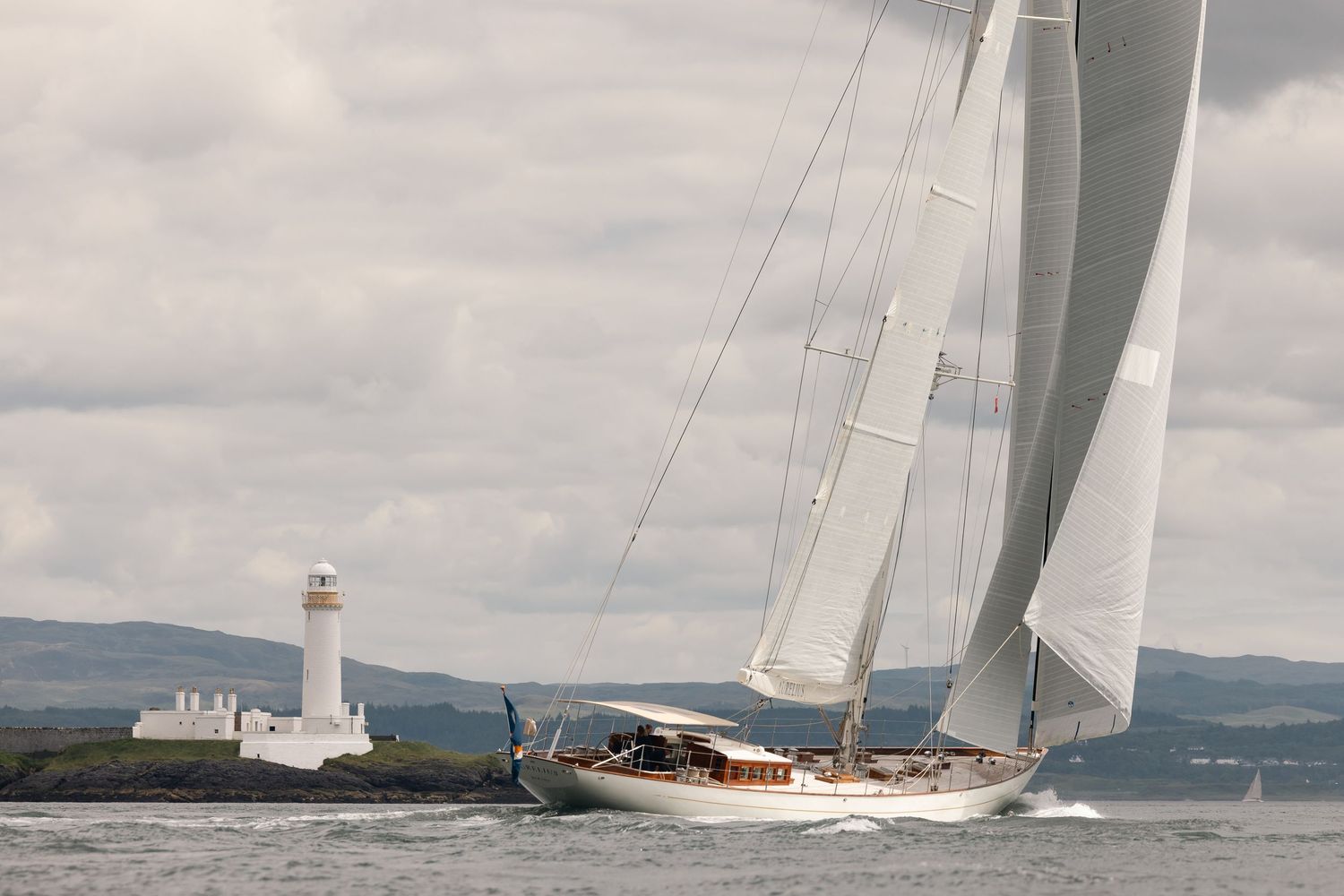 Classic sailboat racing on calm waters with white lighthouse and green hills in background under cloudy skies.