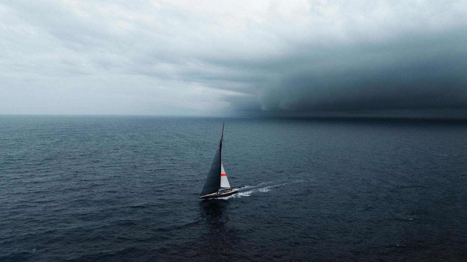Aerial view of a lone sailboat navigating stormy dark ocean waters beneath dramatic threatening storm clouds.