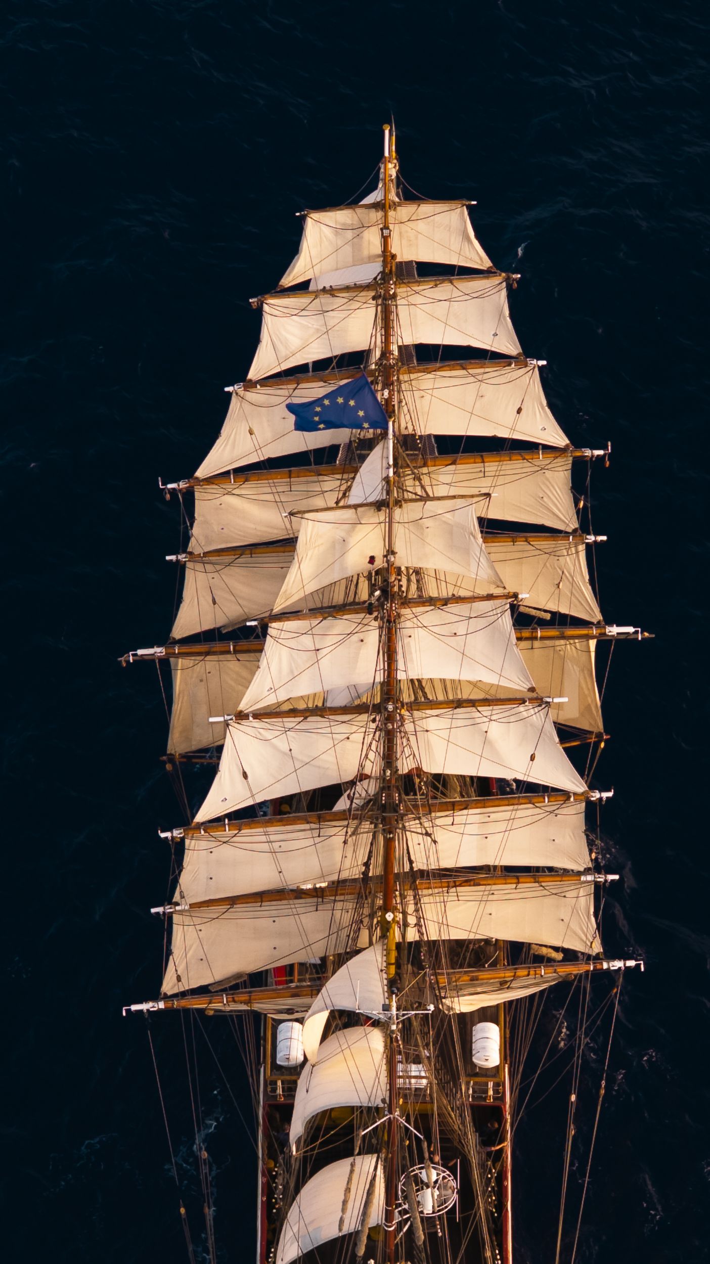 Aerial view of a tall ship with full white sails and EU flag, sailing on dark ocean waters.