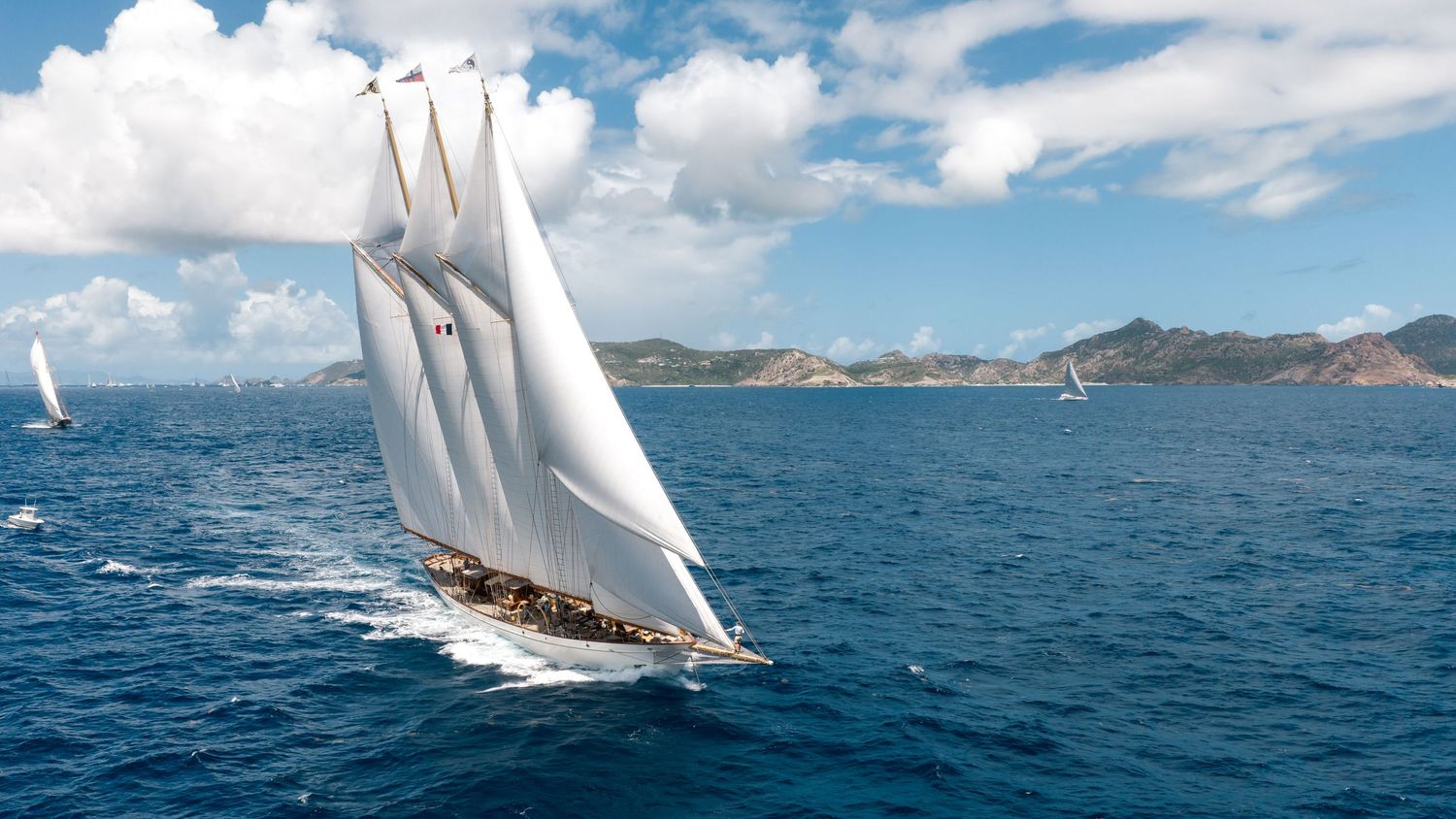 A majestic tall sailing yacht with white sails glides through deep blue ocean waters near a lush green island coastline.