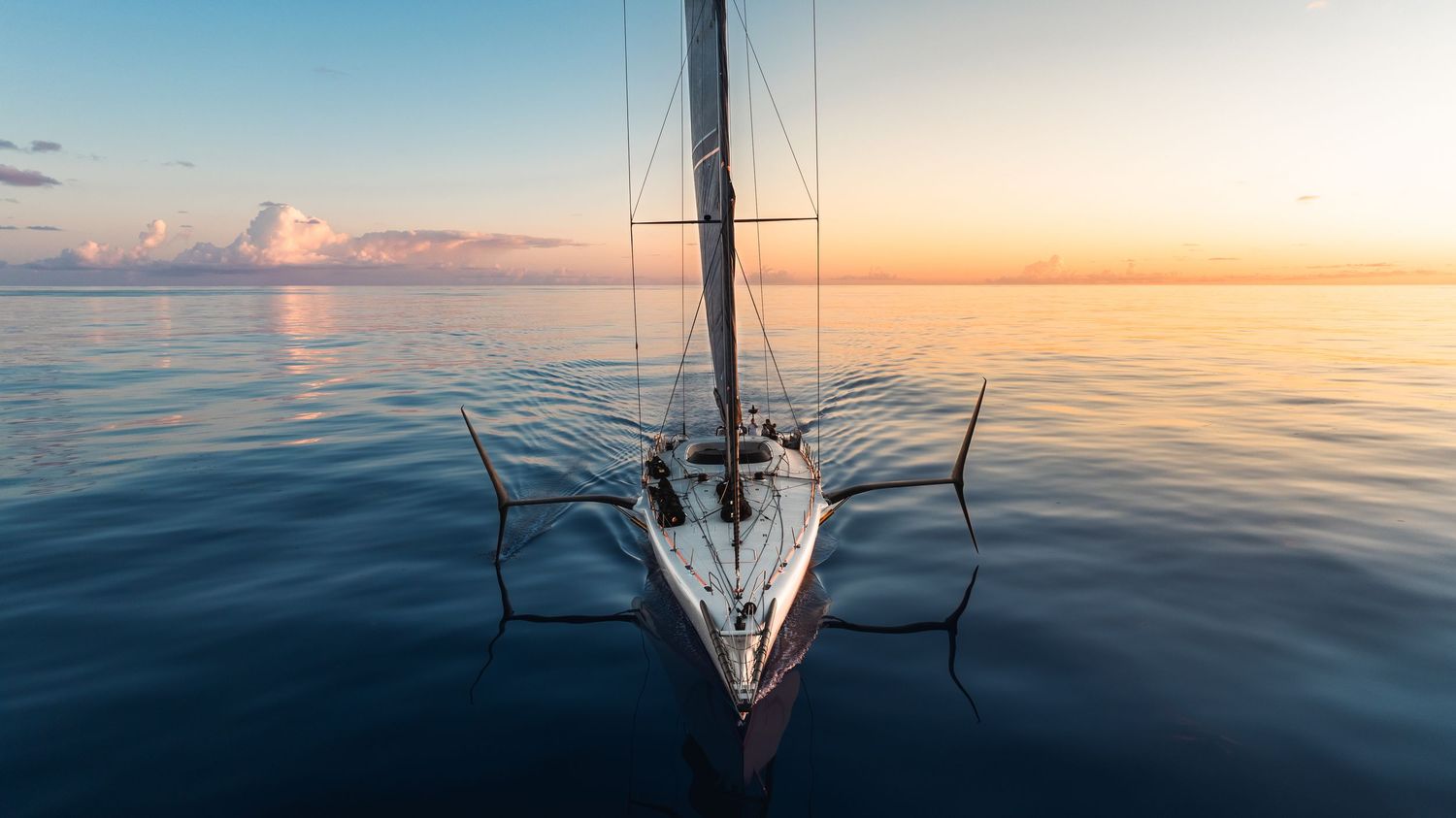 Aerial view of a white sailboat on calm ocean waters at golden sunset with vibrant orange and blue sky reflections.