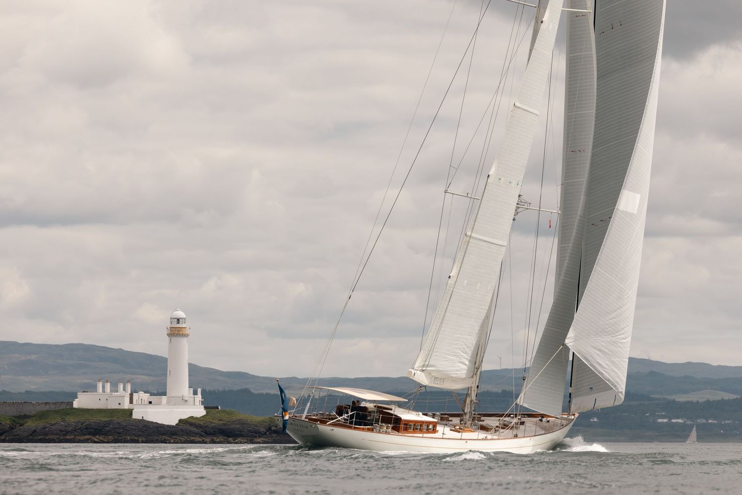 A classic sailboat with white sails glides past a lighthouse on calm waters under a cloudy sky.