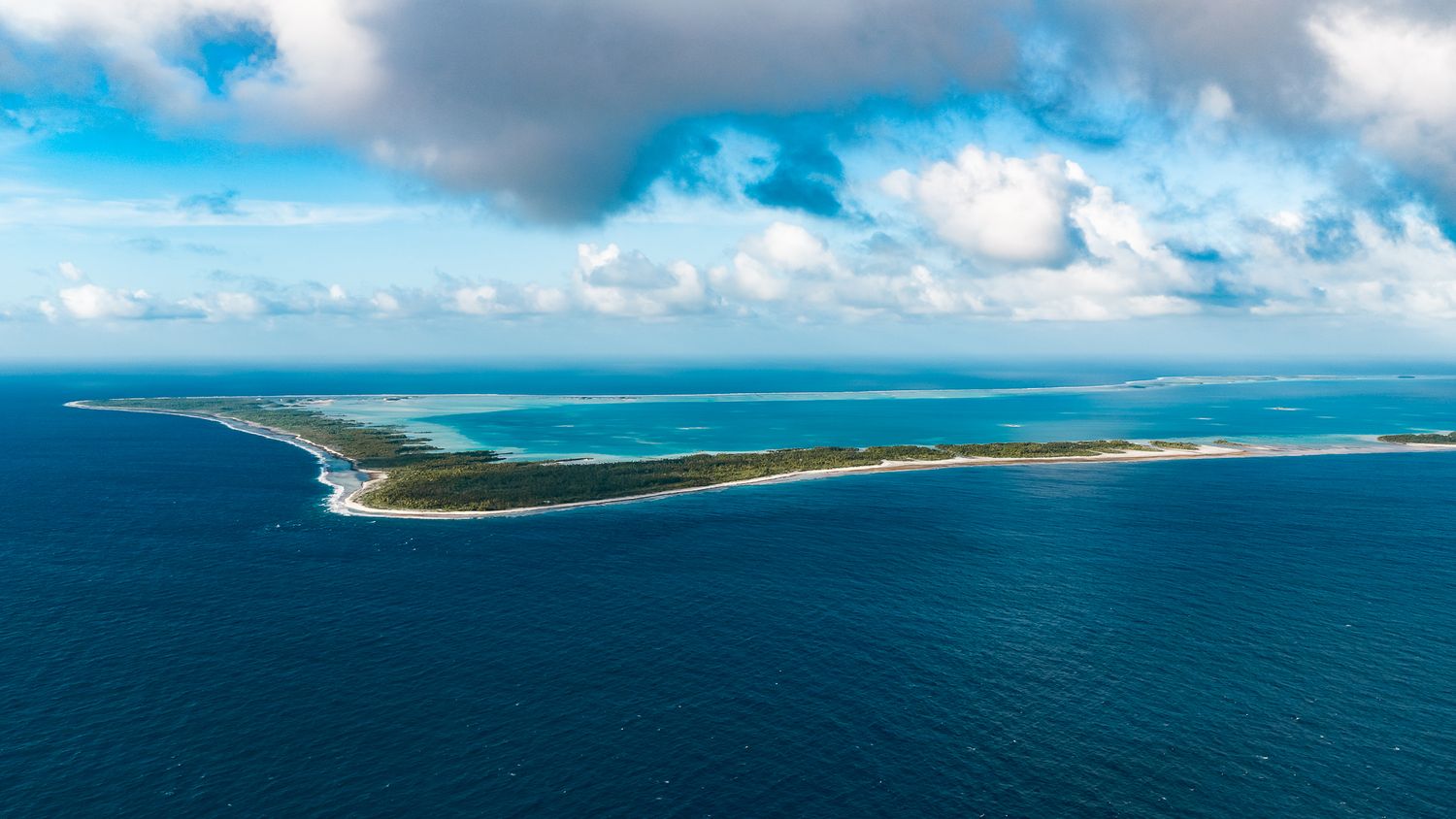 Aerial view of a narrow tropical island surrounded by turquoise and deep blue ocean waters under a partly cloudy sky.