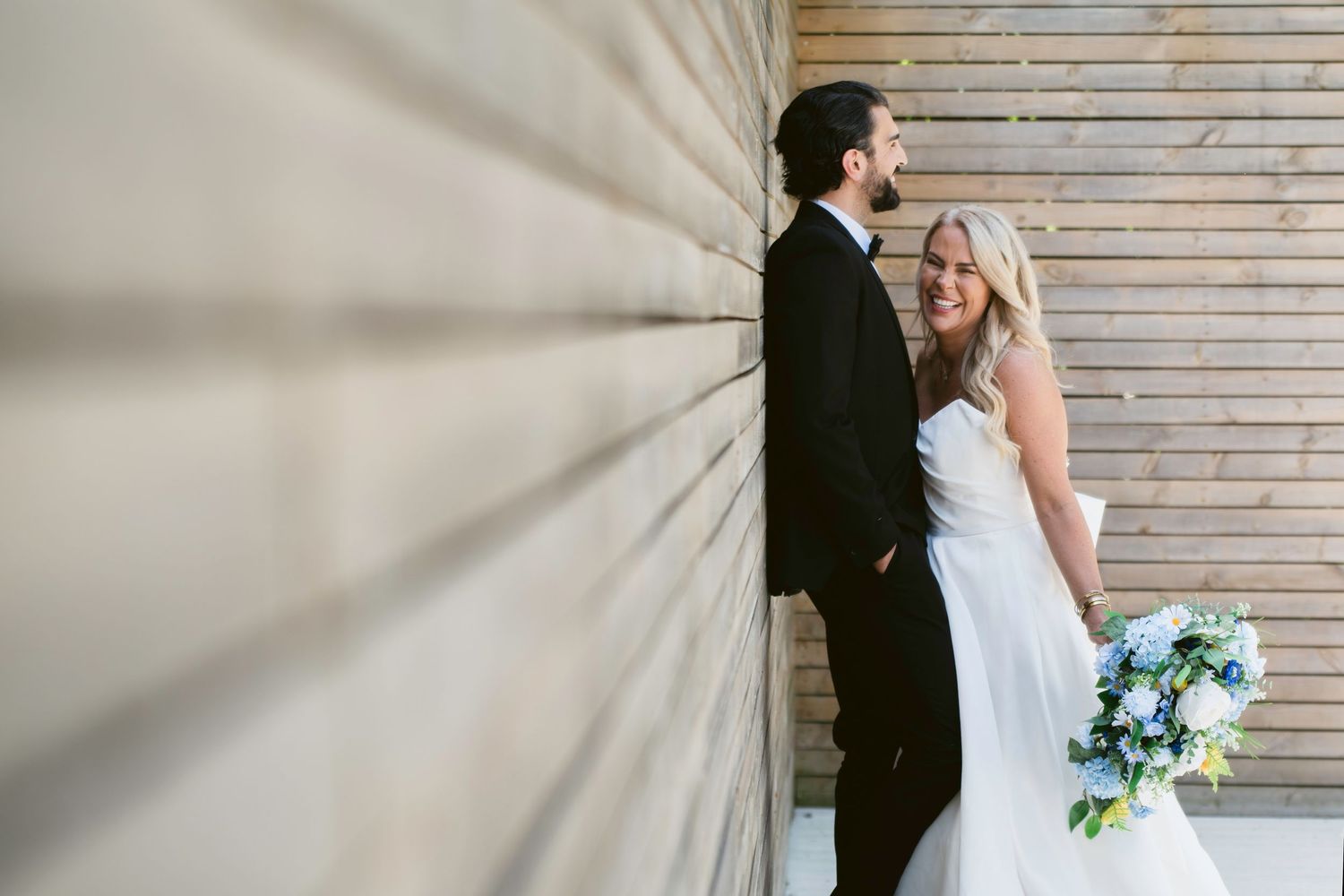 Bride in white gown holding blue bouquet smiles at groom in black tux leaning against wooden wall.