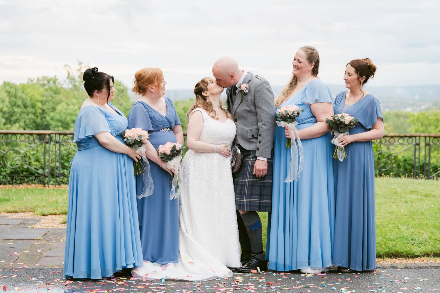 Bride and groom kissing surrounded by four bridesmaids in blue dresses holding bouquets at an outdoor wedding ceremony.