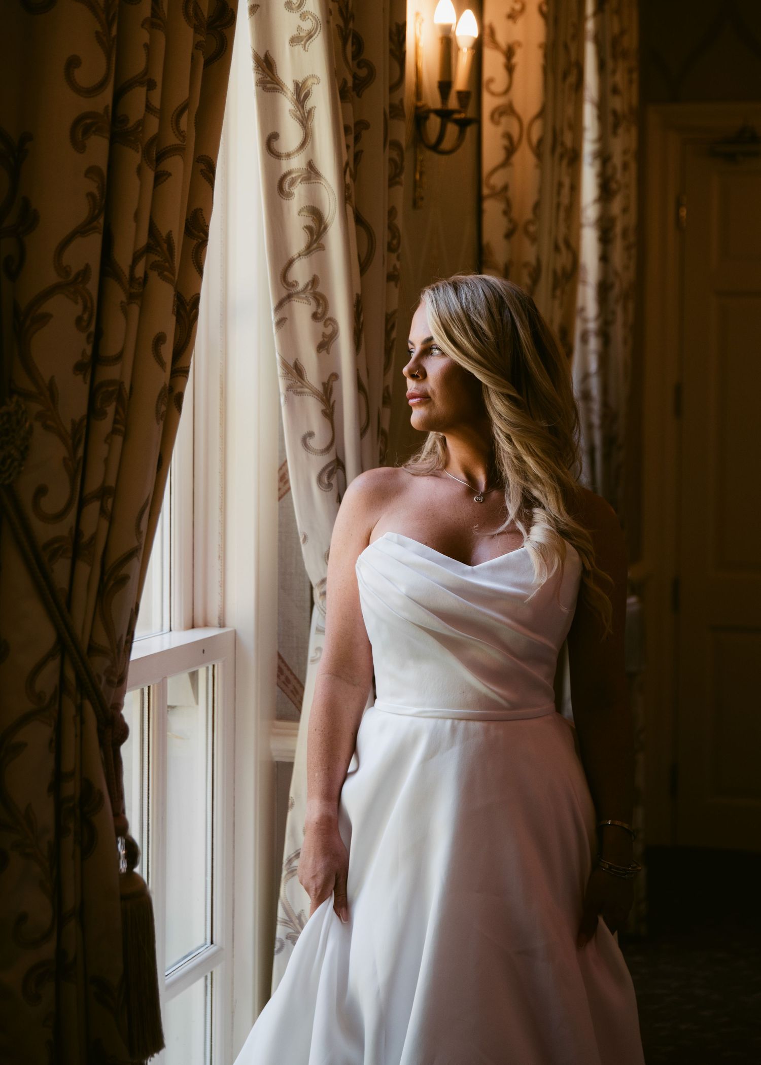 Bride in strapless white wedding gown stands by ornate window, gazing out in elegant decorated room.