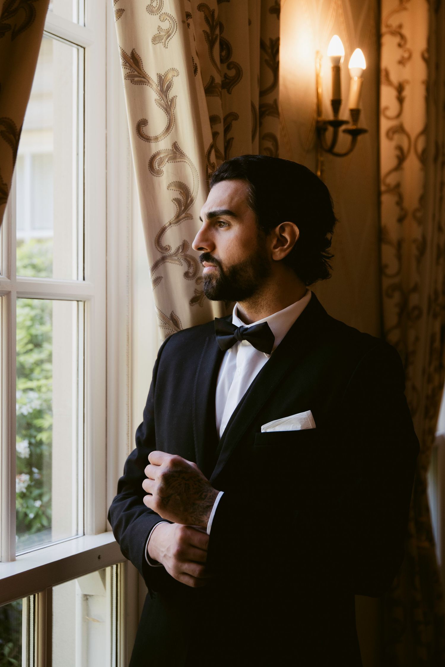 A dapper groom in a black tuxedo and bow tie gazes thoughtfully out a window in an elegant room.