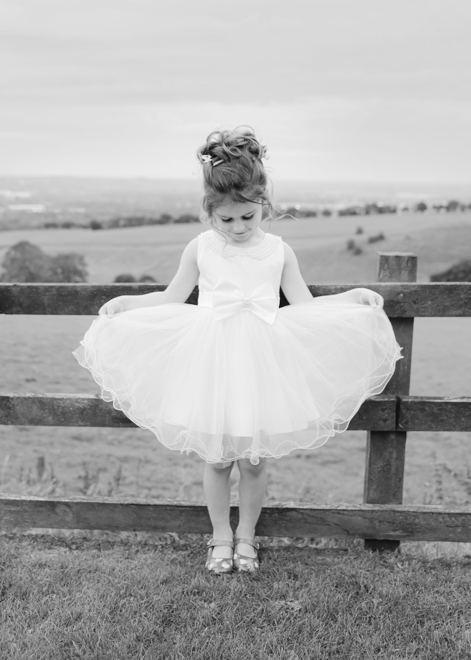 A young flower girl in a white tulle dress holds out her skirt while standing by a wooden fence in a rural landscape.