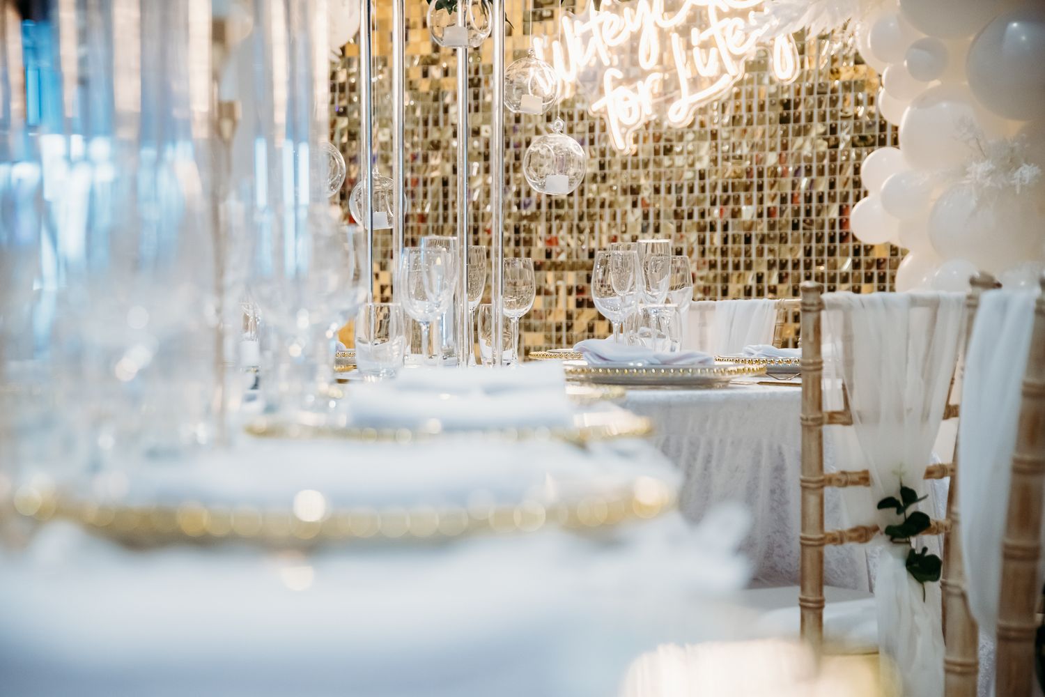 Elegant wedding reception table setup with gold and white decor, crystal glasses, and a shimmering gold mosaic backdrop.