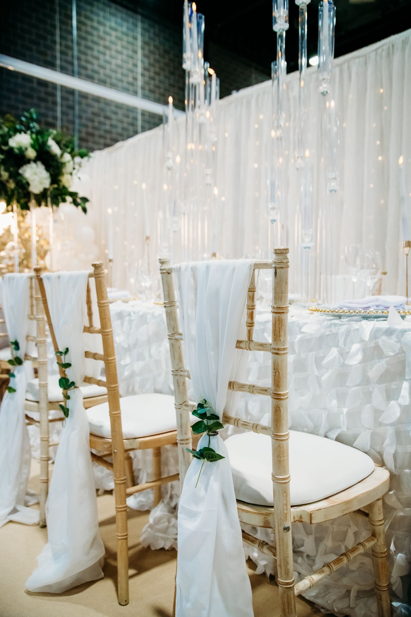 Elegant wedding reception chairs with white fabric sashes and green accents, gold chiavari chairs, candles, and white floral decor.