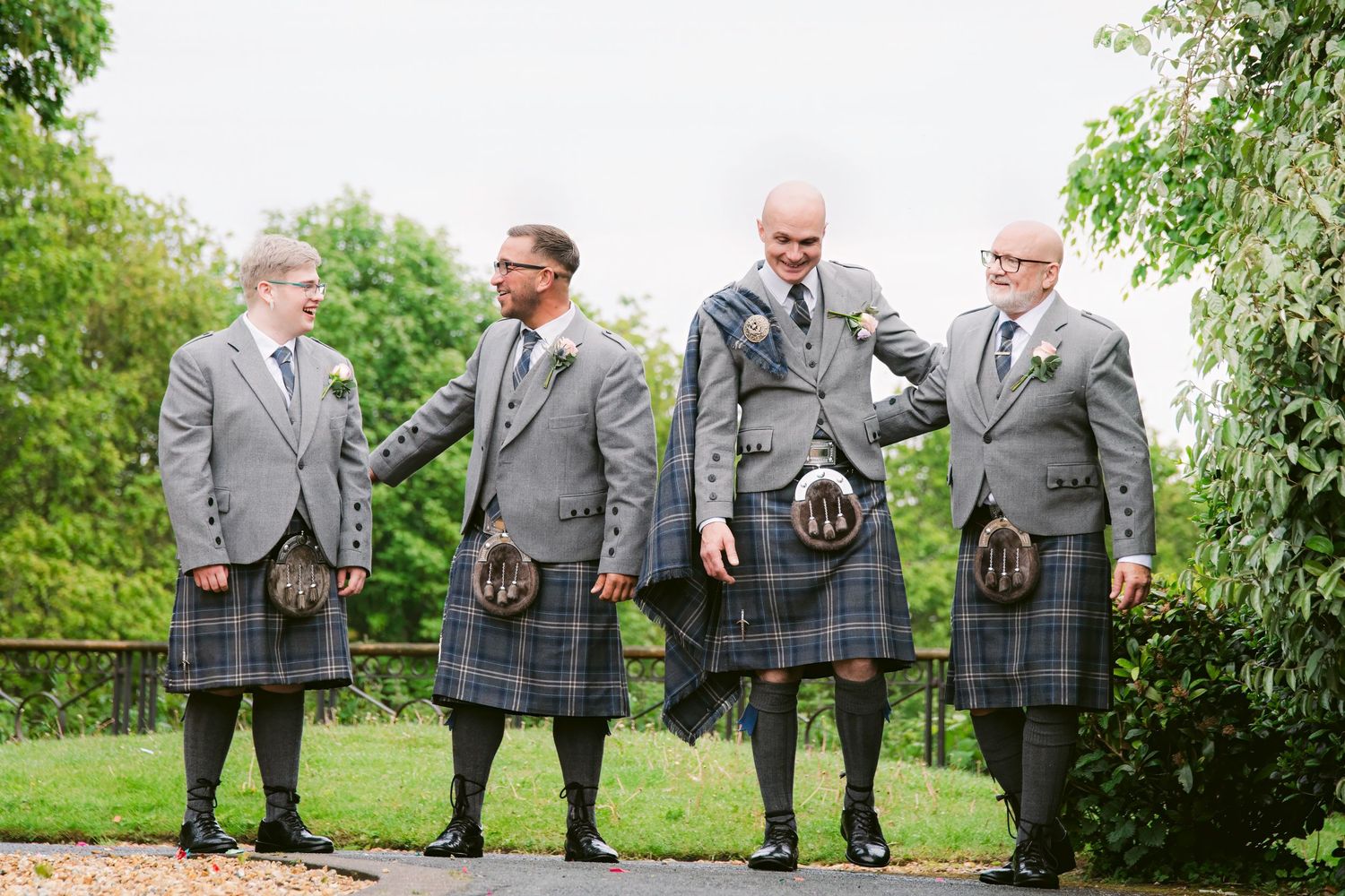 Four men in traditional Scottish kilts and grey jackets stand outdoors on a path surrounded by lush green trees.