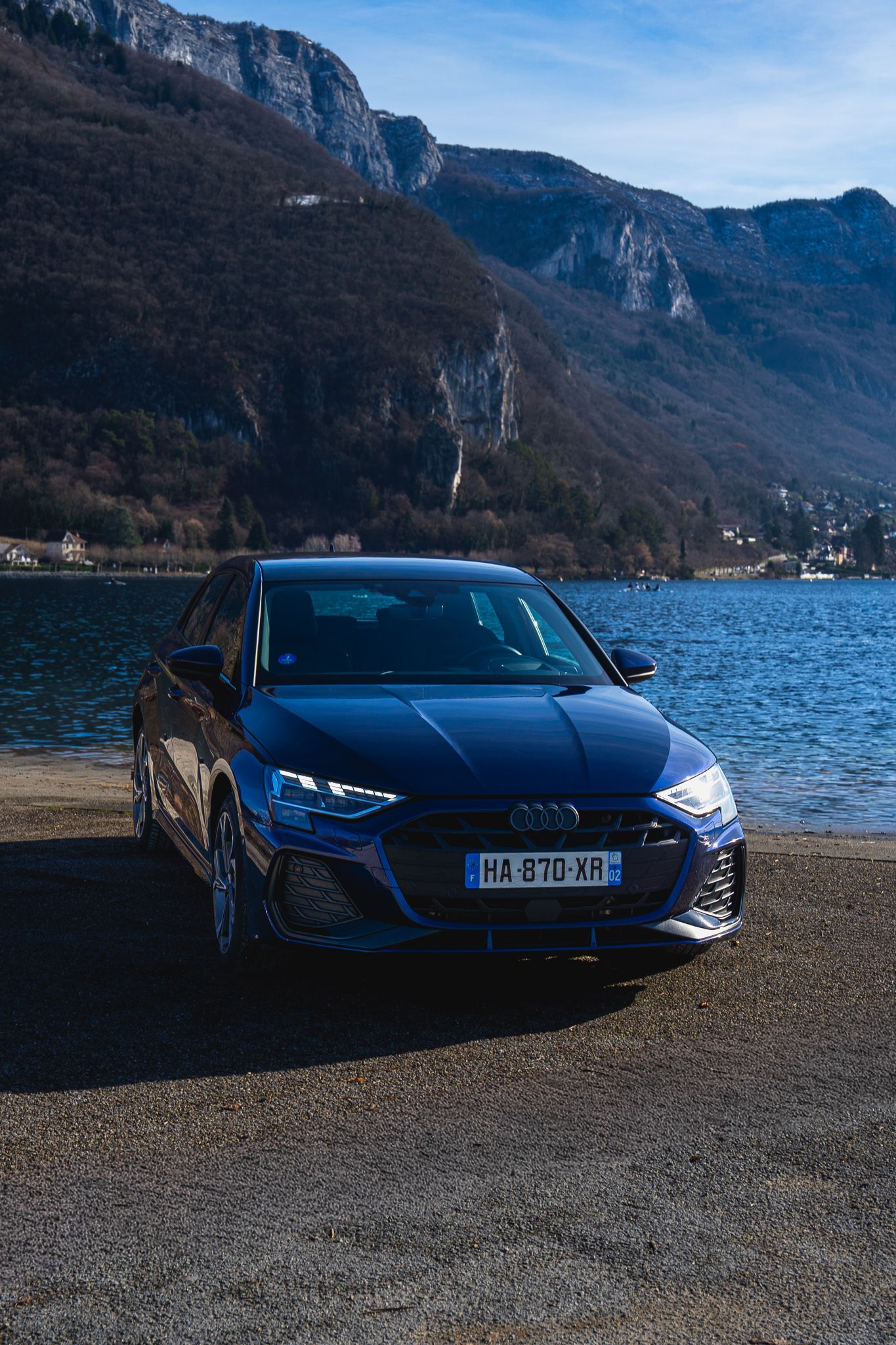 A sleek blue Lamborghini Aventador parked along a scenic mountain lake with dramatic cliffs in the background.