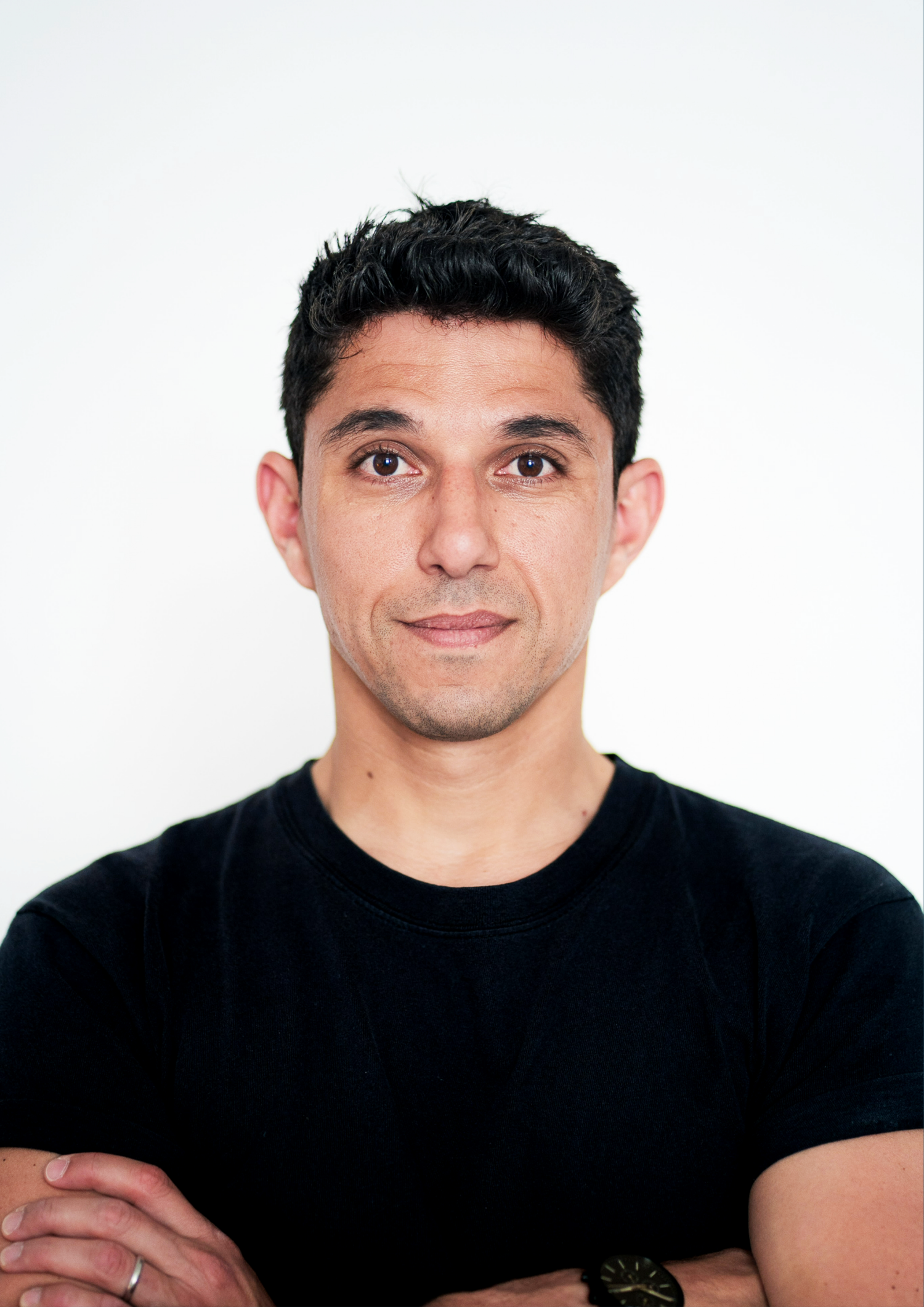 A man with short dark hair wearing a black t-shirt stands with arms crossed against a white background.
