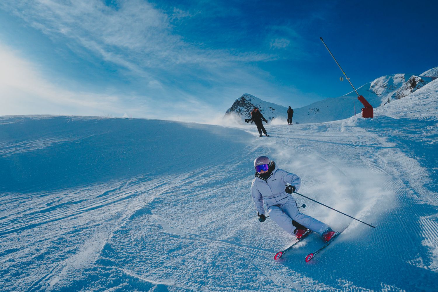 Skier performs dynamic turns on a pristine snow-covered mountain slope against bright blue winter sky.