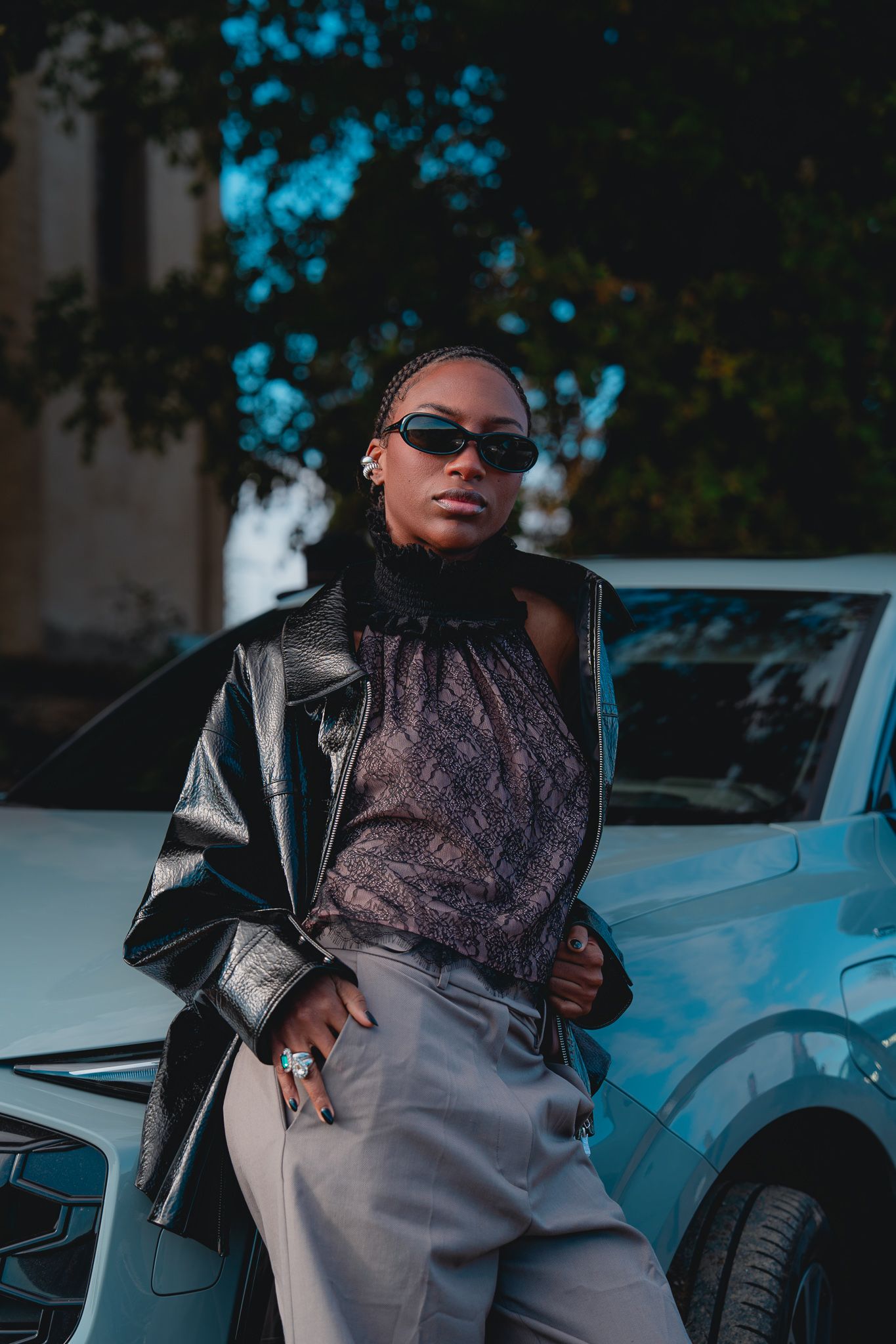 Fashion model in sunglasses and leather jacket poses confidently next to luxury car in urban setting.