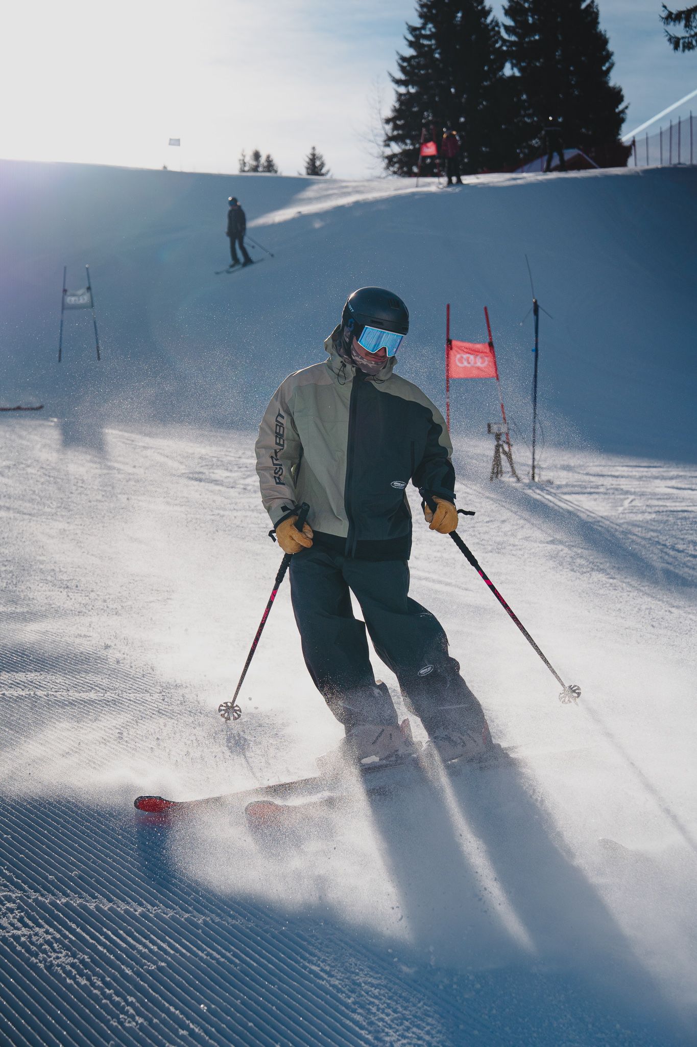 A skier carves through fresh powder snow creating a dramatic spray against a blue winter sky on a groomed slope.