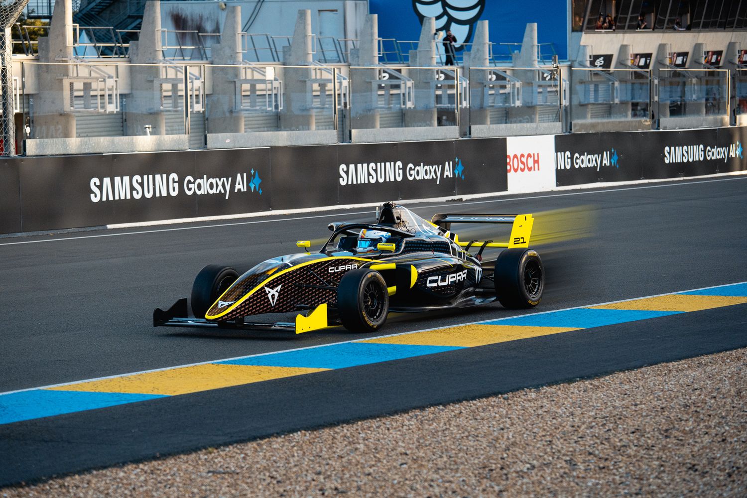 Formula racing car speeds along a race track with blue barriers and Samsung Galaxy branding visible on track walls.