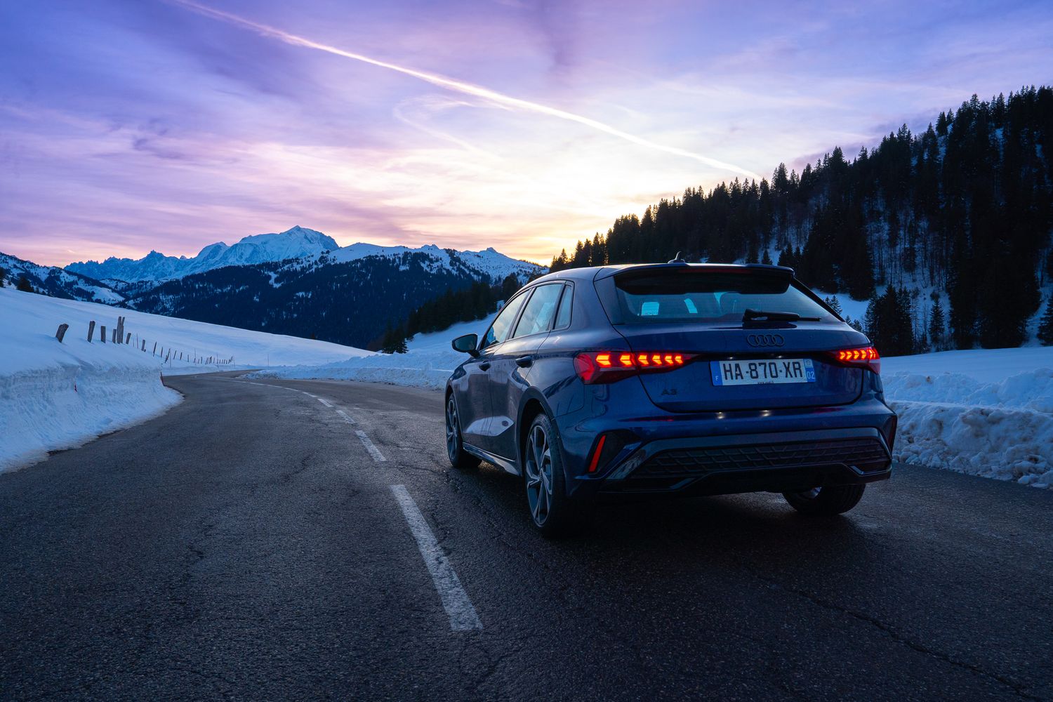 Blue Audi RS3 sports car parked on snowy mountain road at sunset with dramatic purple sky and mountain peaks.