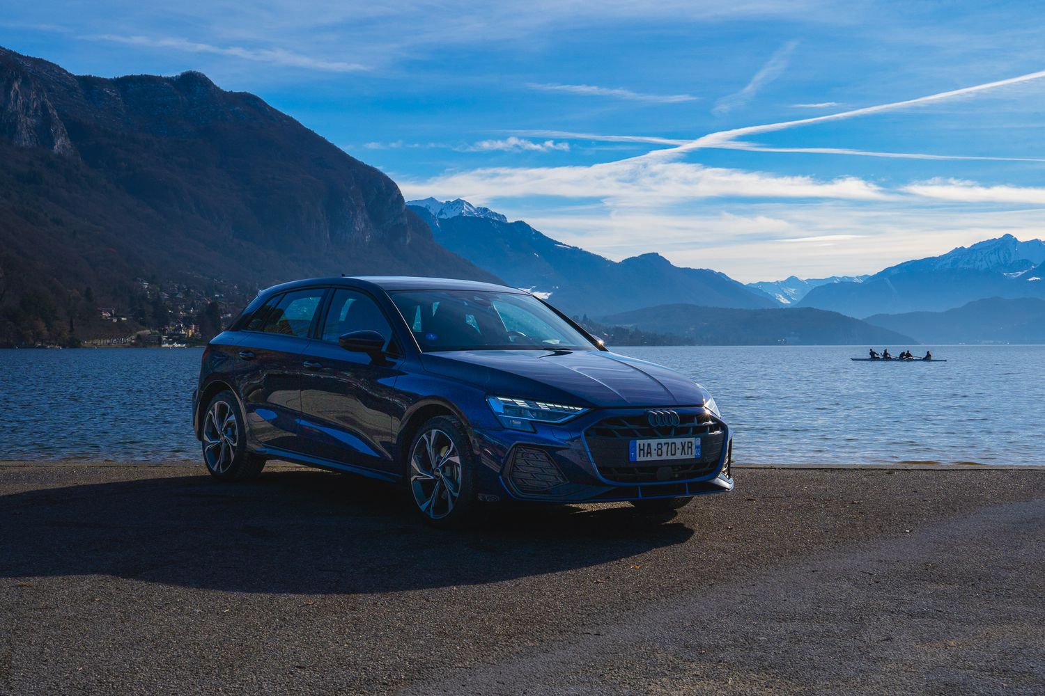 Blue McLaren supercar parked along scenic mountain lake with dramatic cloudy sky and reflective water.