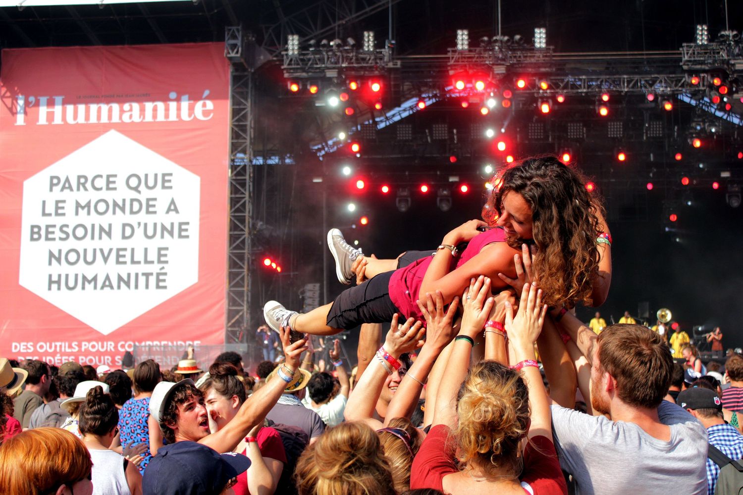 Concert crowd carries a performer during an outdoor music festival with red lights and L'Humanite banner in background.