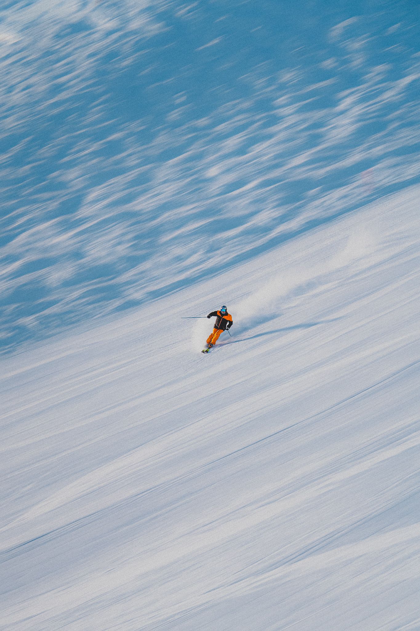 A lone snowboarder carves down a pristine snowy slope against a bright blue sky background.