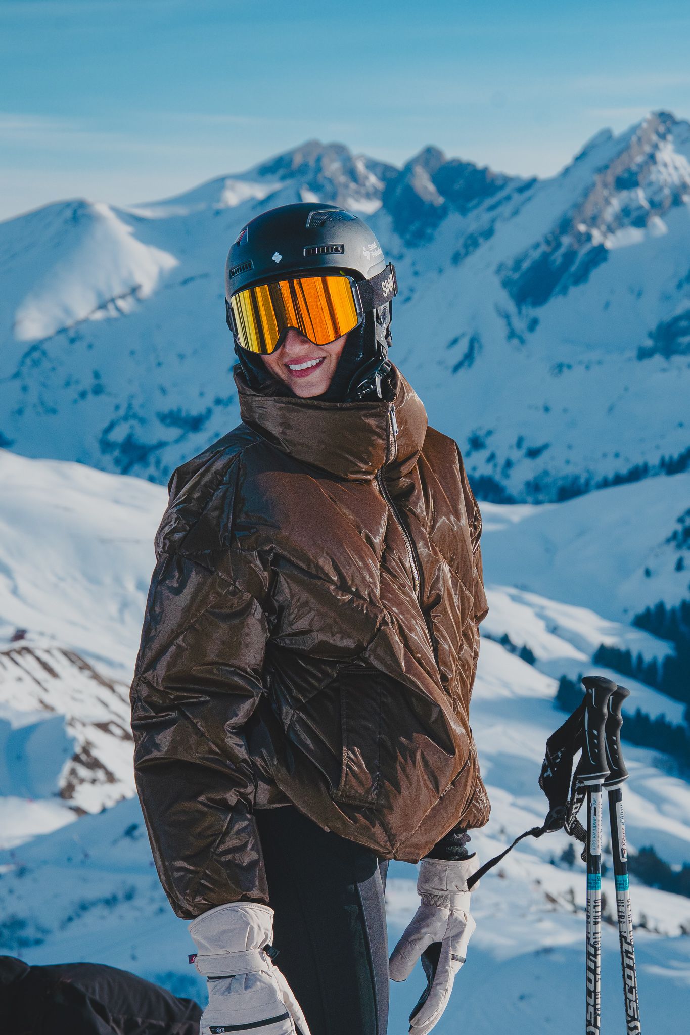 Skier in brown winter jacket and goggles stands against snowy mountain peaks on a bright winter day.