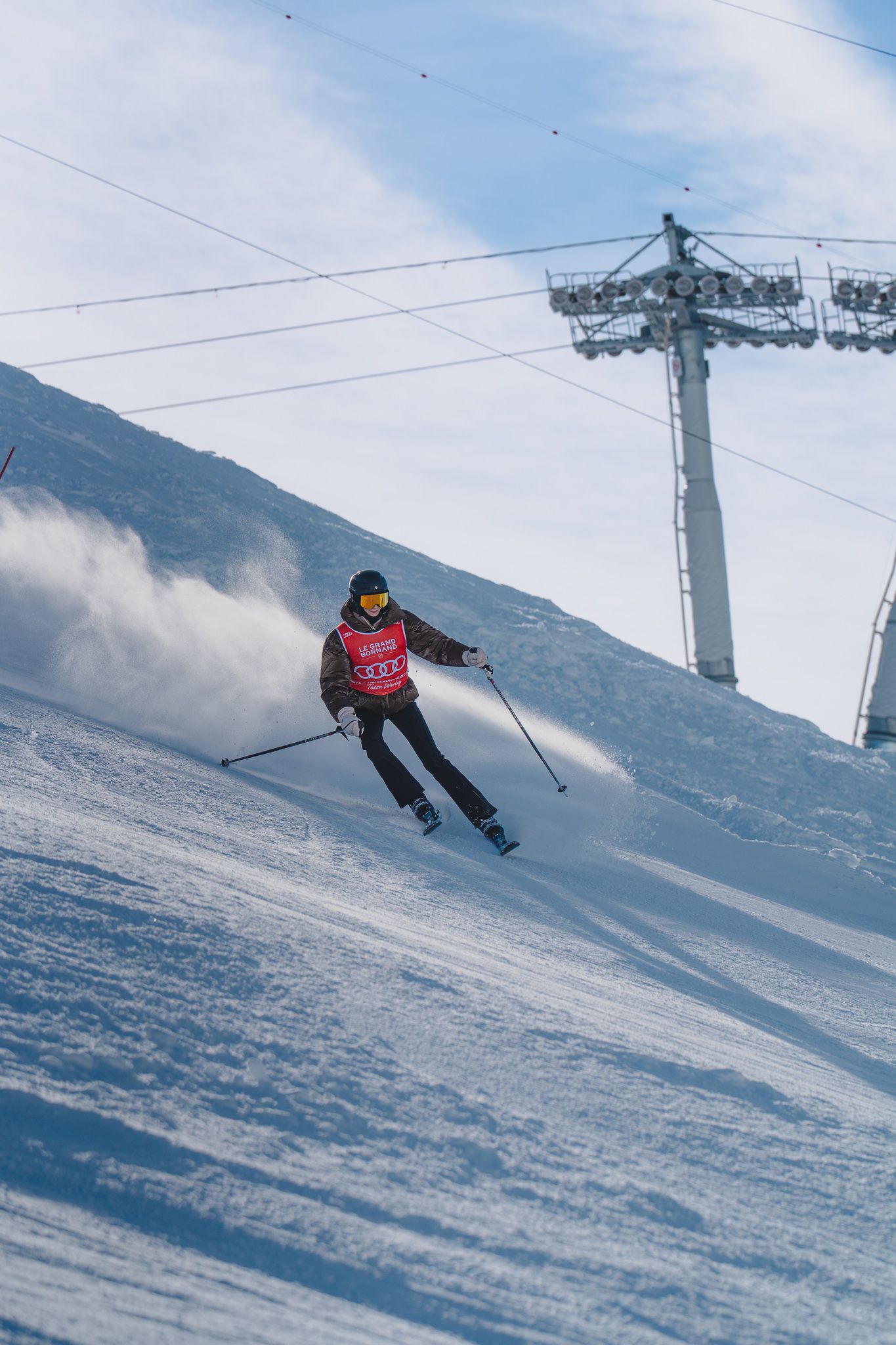 A skier carves through fresh powder snow on a steep mountain slope with ski lift towers visible in the background.