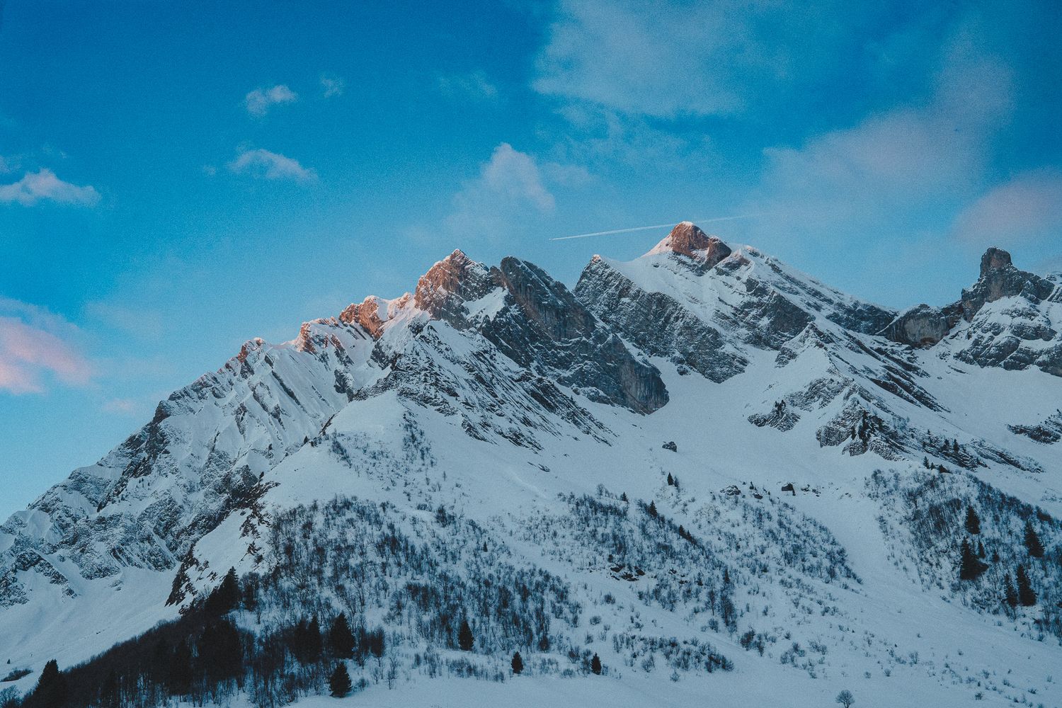 A panoramic view of snow-covered mountain peaks against a bright blue sky with wispy clouds.