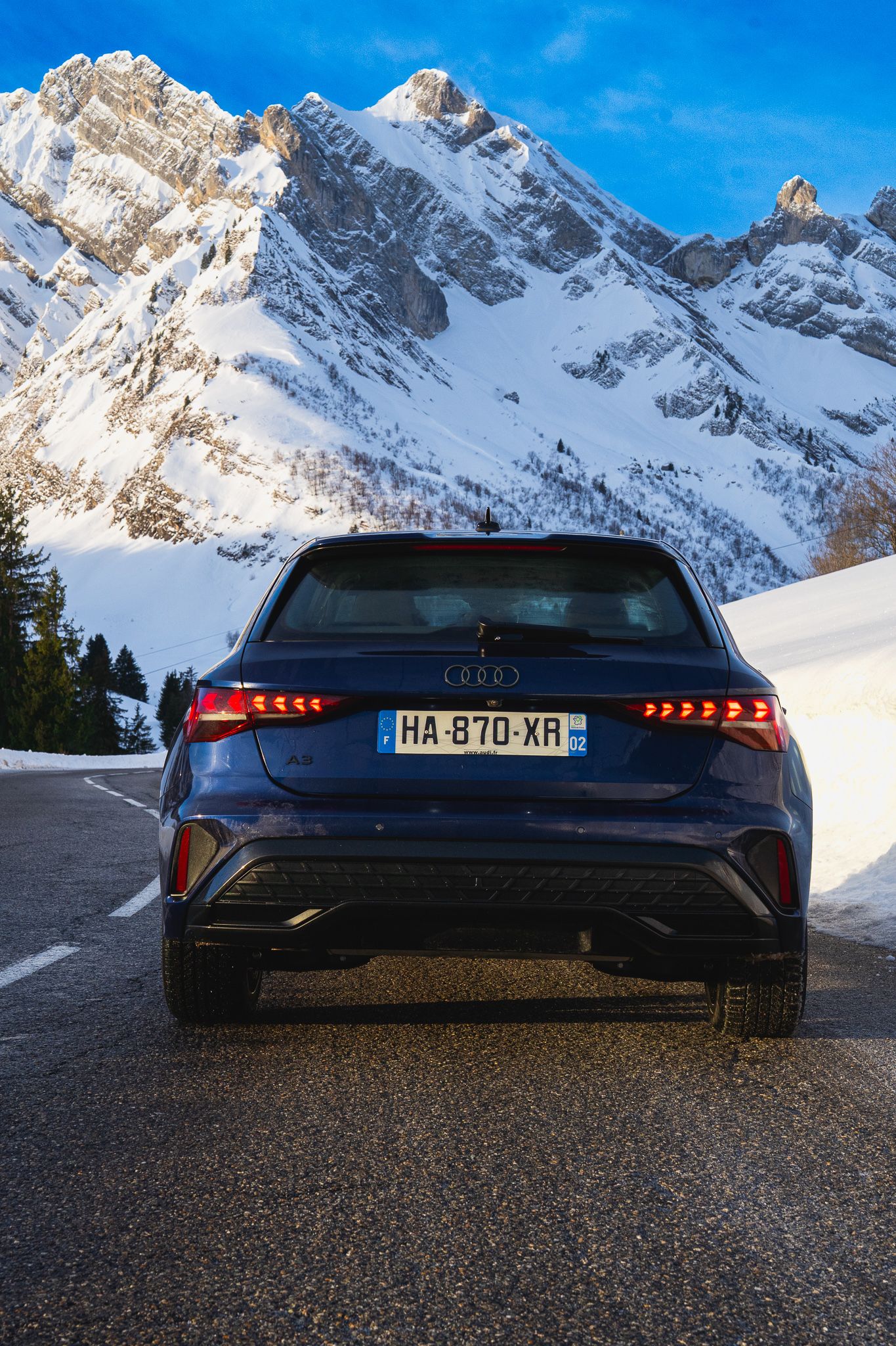 Dark luxury car parked on mountain road with snowy Alpine peaks and blue sky in background.