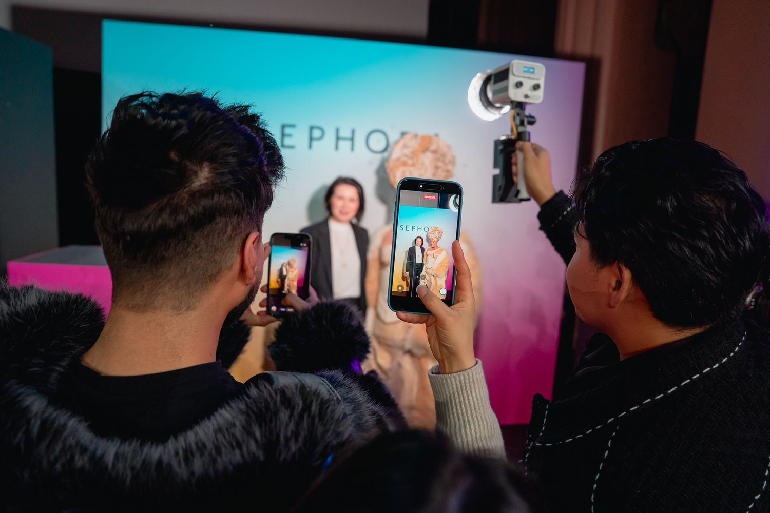 People gather in a dark room taking photos with their phones at an event with colorful lighting.
