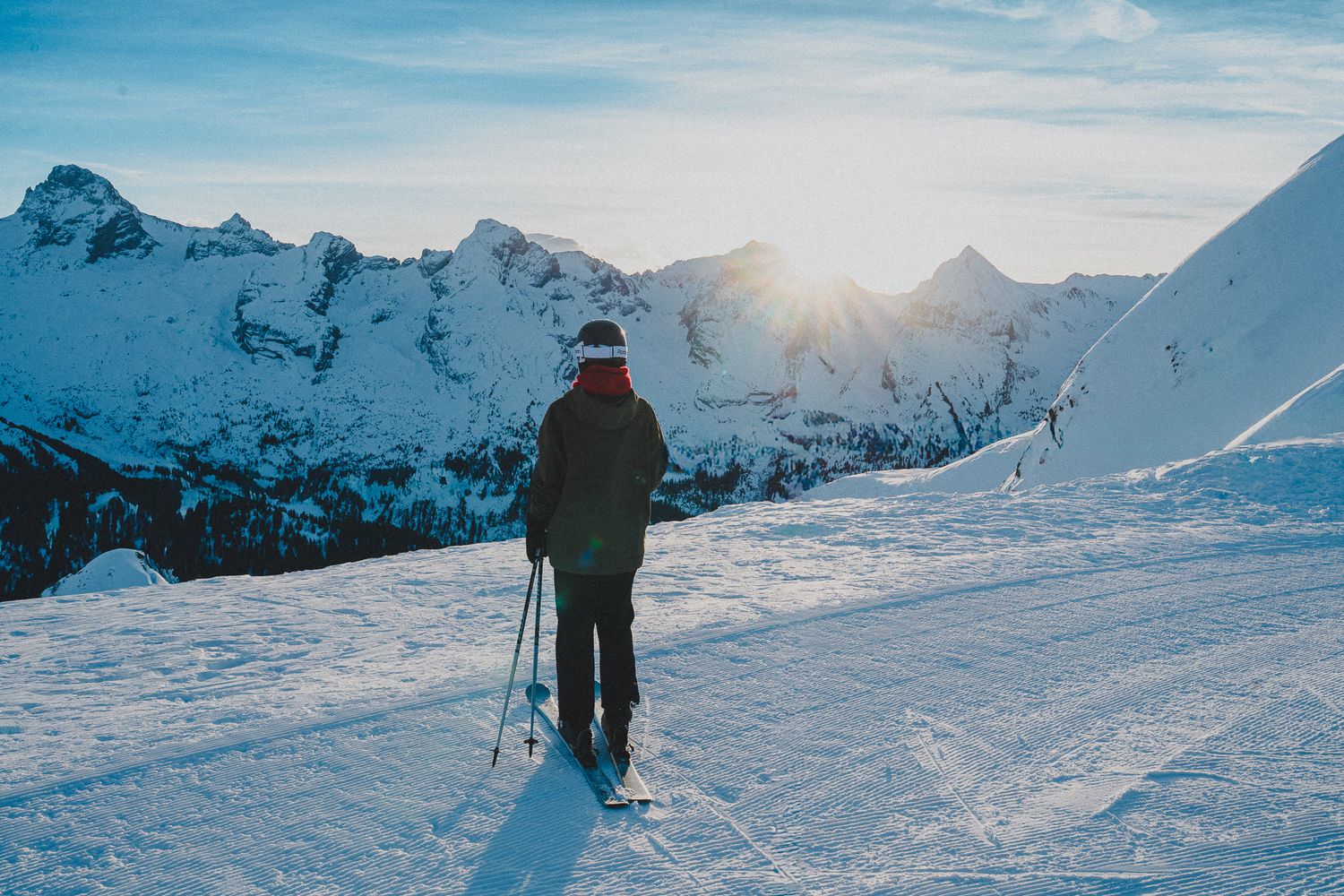 Skiers traverse a snowy mountain slope with dramatic snow-capped peaks and bright sunlight in the background.
