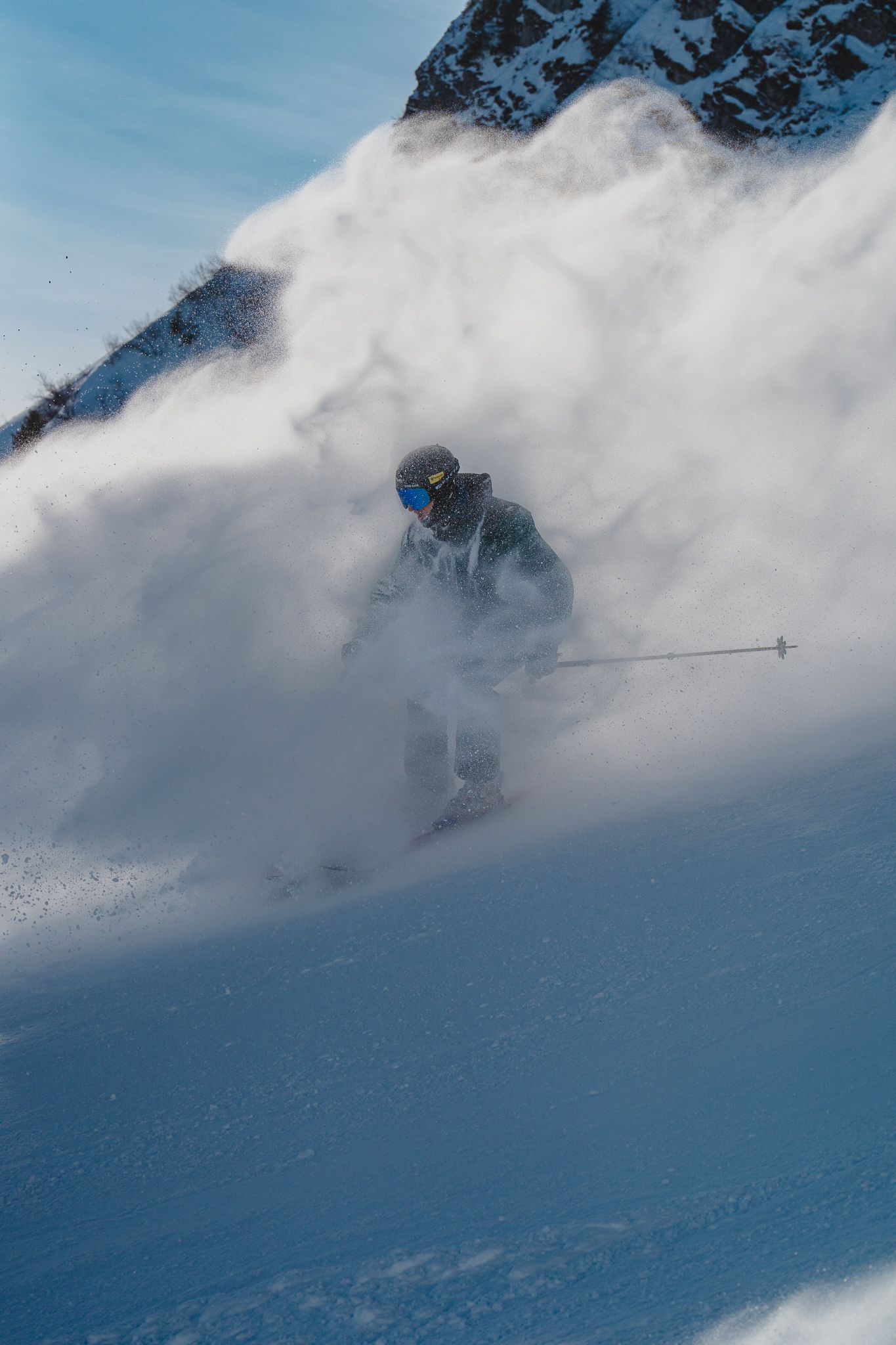 A skier carves through deep powder snow creating a dramatic spray while descending a steep mountain slope in misty conditions.