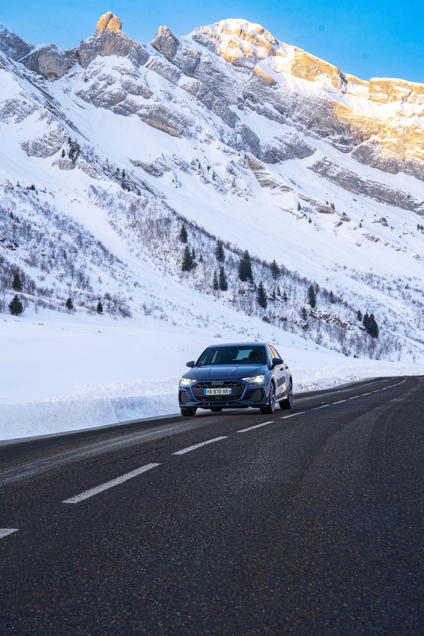 A black sports car drives on a winding mountain road surrounded by snow-covered peaks and clear blue winter skies.